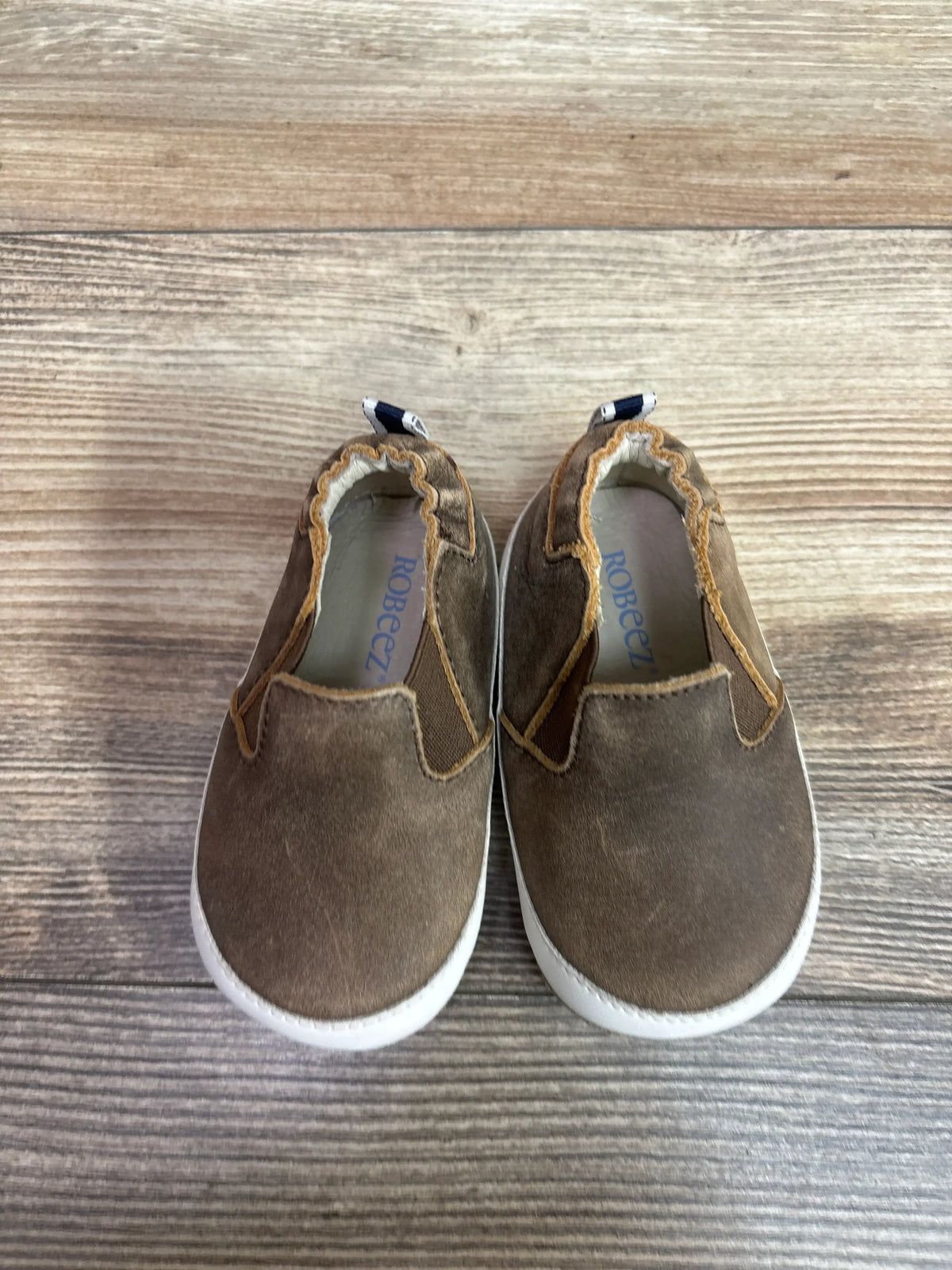 Top view of Robeez Lenny Loafer crib shoes showcasing brown uppers and white soles on a wood-grain background