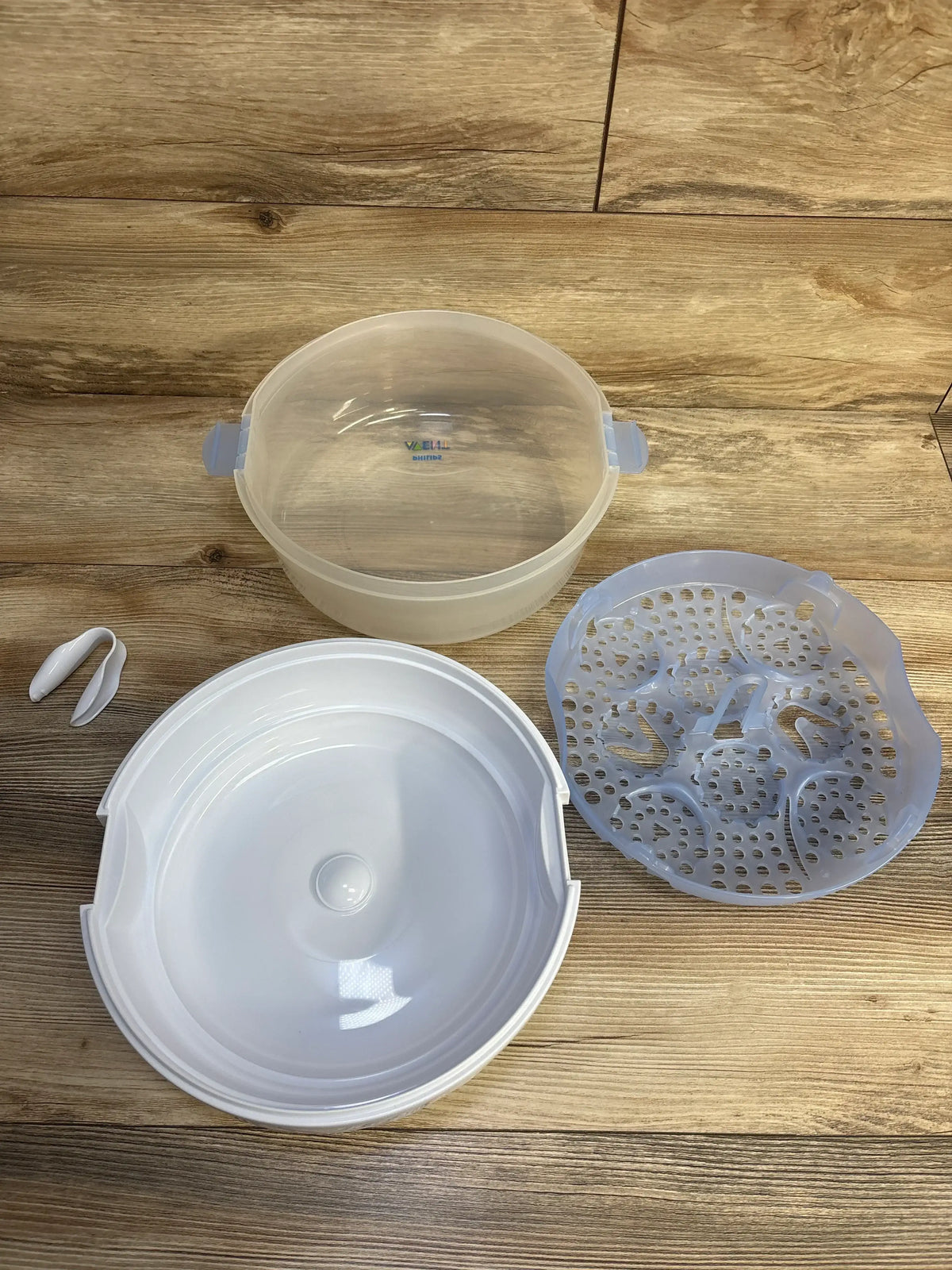 Top down view of AVENT microwave steam sterilizer parts: lid, perforated tray, water base and tongs on wood