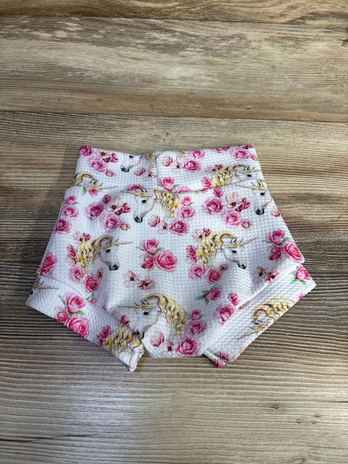 Top view of white baby bloomers (12m) showing unicorn and pink rose print on textured knit, on wood background