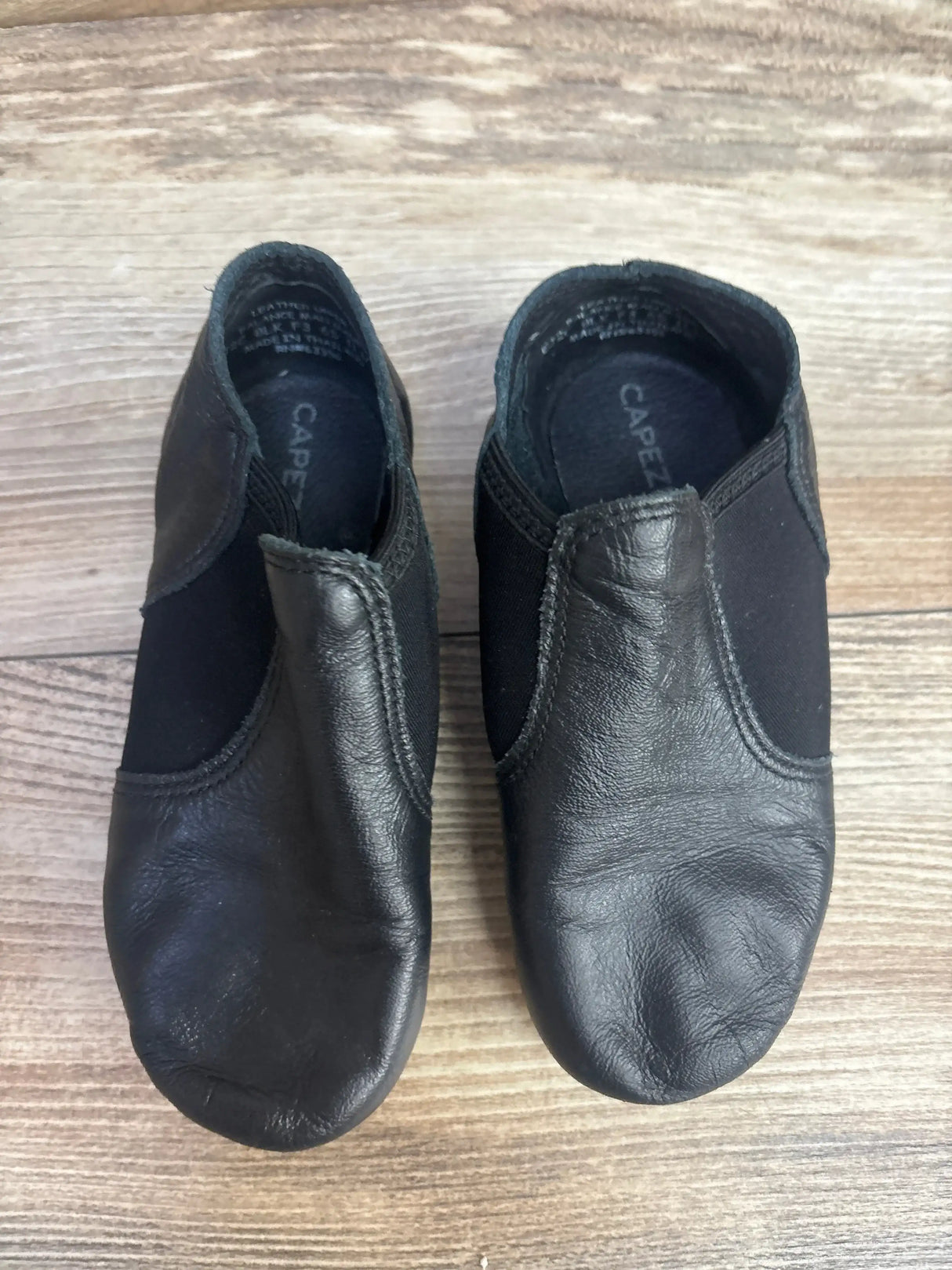 Top-down view of Capezio EJ2C girls’ black jazz slip-on shoes, leather front with elastic side panels, size 11.5C on wood floor