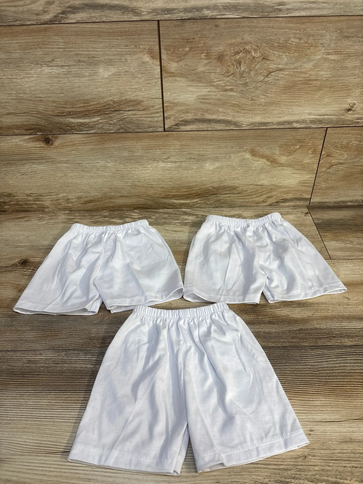 Three pairs of white kids' shorts with elastic waistbands on wood floor, ideal for baby and toddler clothing resale