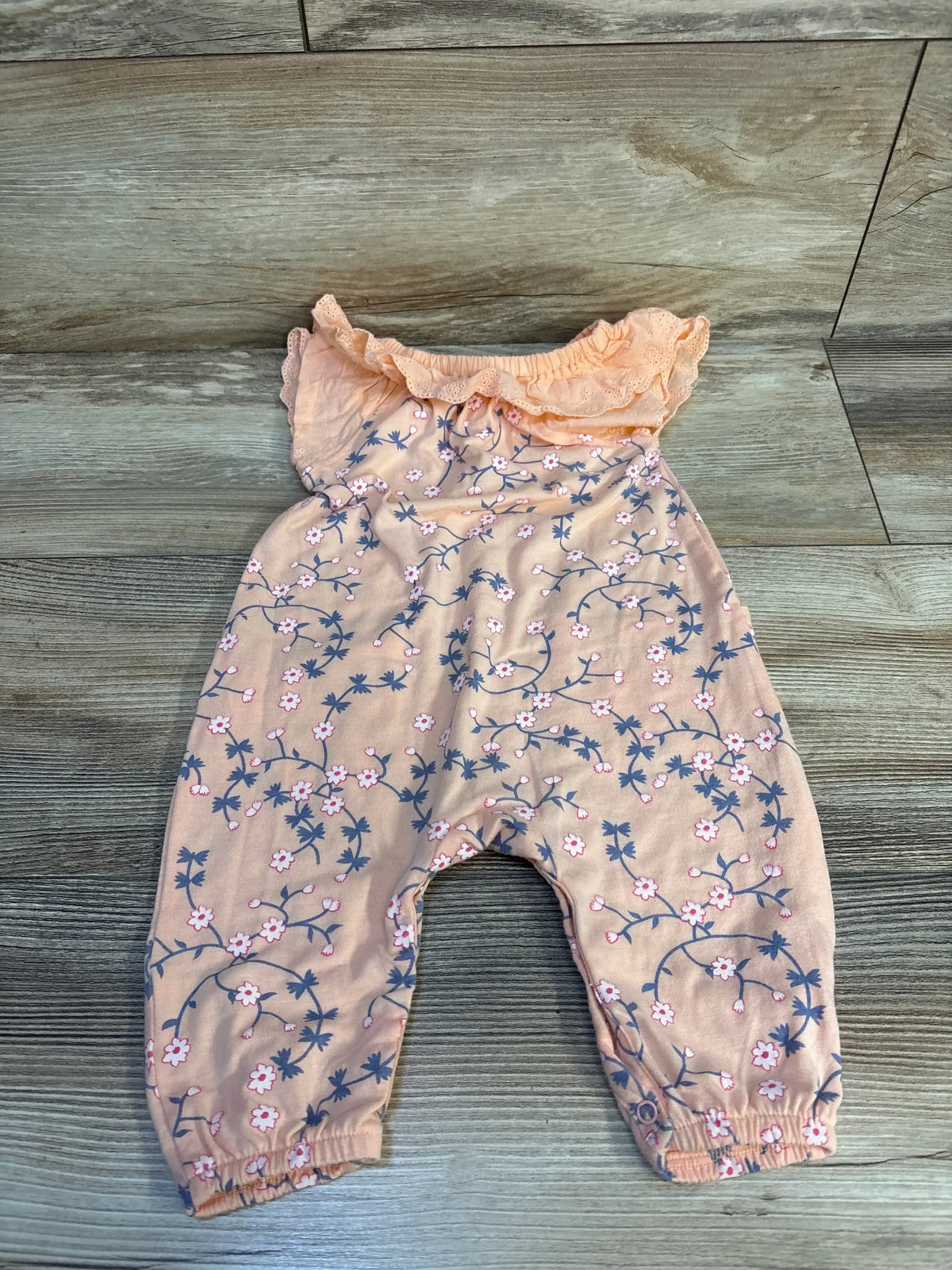 Flat lay of Jessica Simpson peach floral jumpsuit size 3-6M showcasing ruffled neckline, elasticized ankle cuffs, and all-over pink and navy flower print on wooden floor background