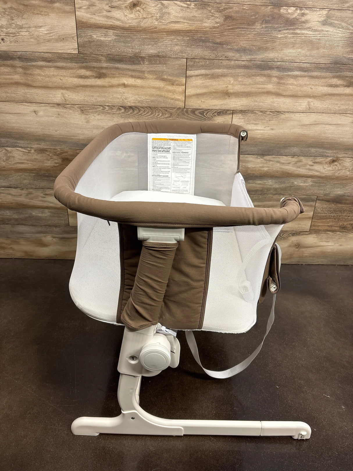 Angled view of Baby Delight Beside Me Dreamer bedside bassinet showcasing mesh side panels, padded mocha rim, and white adjustable support leg against wood-paneled background