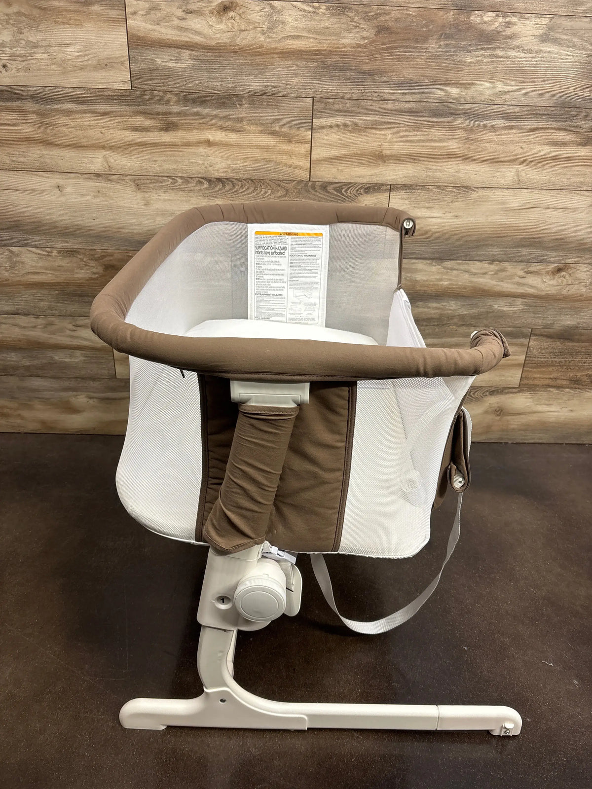 Angled view of Baby Delight Beside Me Dreamer bedside bassinet showcasing mesh side panels, padded mocha rim, and white adjustable support leg against wood-paneled background