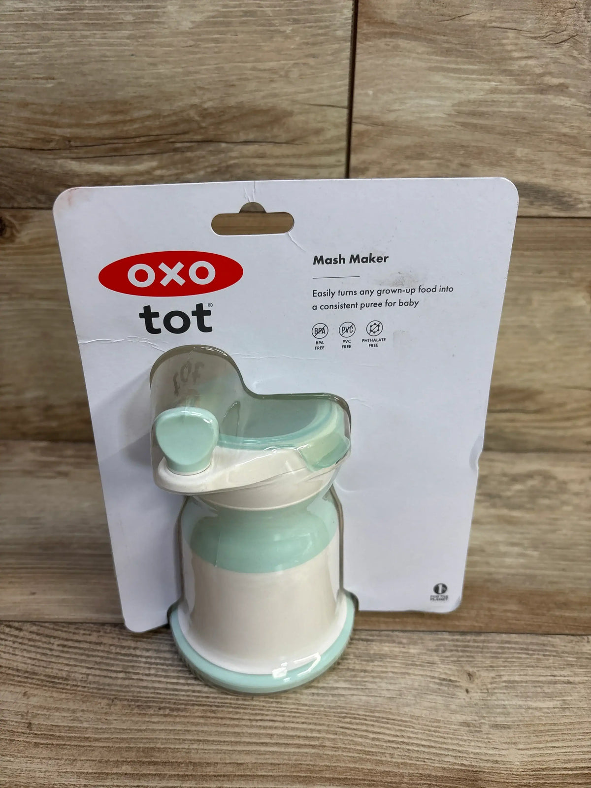 Angled front view of OXO Tot Mash Maker baby food mill showing mint opal plunger and white base in original packaging