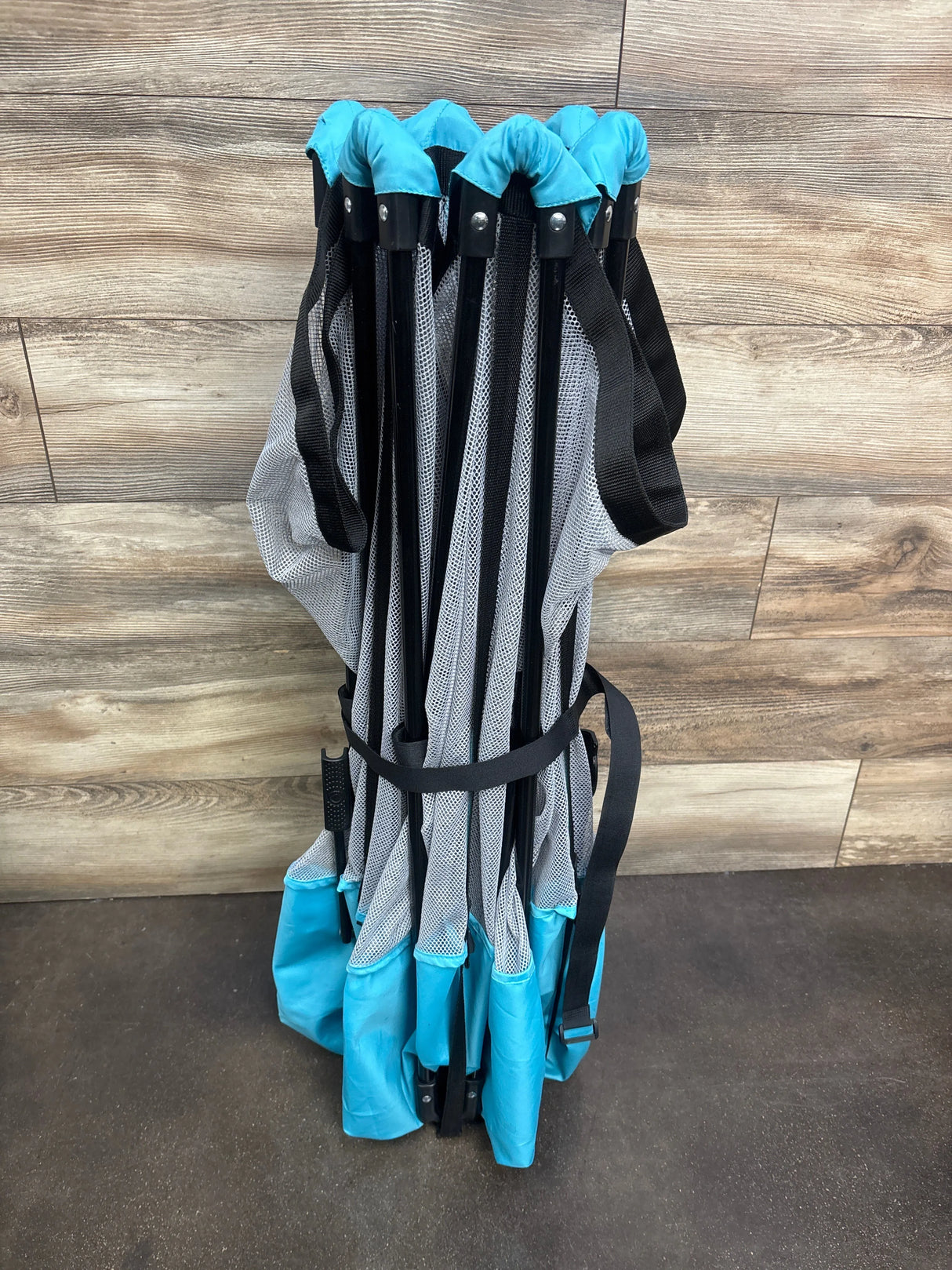 Folded Regalo My Play 6-panel play yard in teal showing compact storage, black frame and mesh panels on wood floor