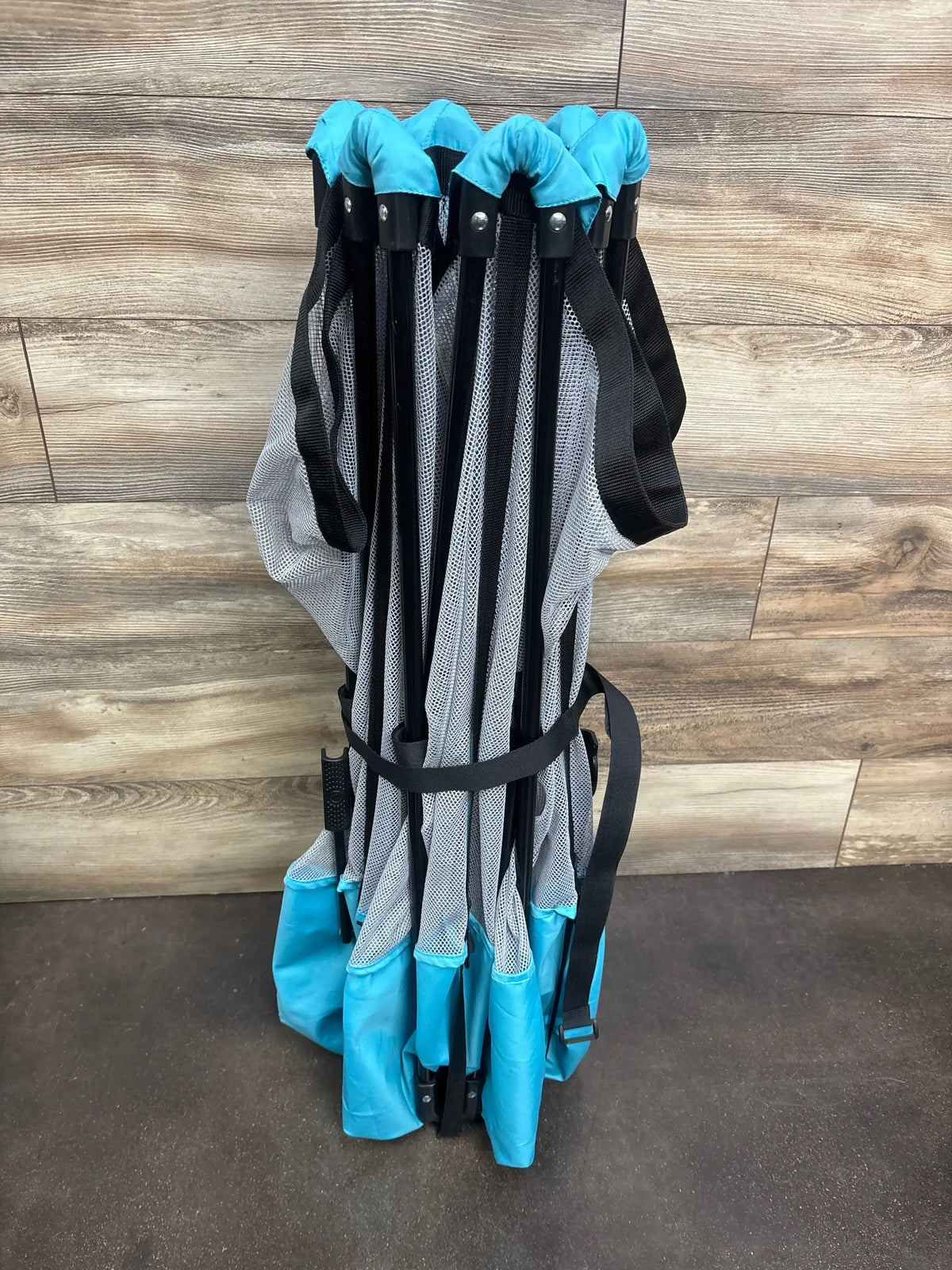Folded Regalo My Play 6-panel play yard in teal showing compact storage, black frame and mesh panels on wood floor