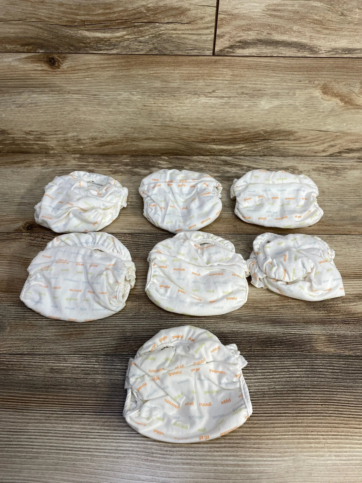 Top-down view of seven gDiapers newborn cloth diapers in ivory with orange and green word print on wood background