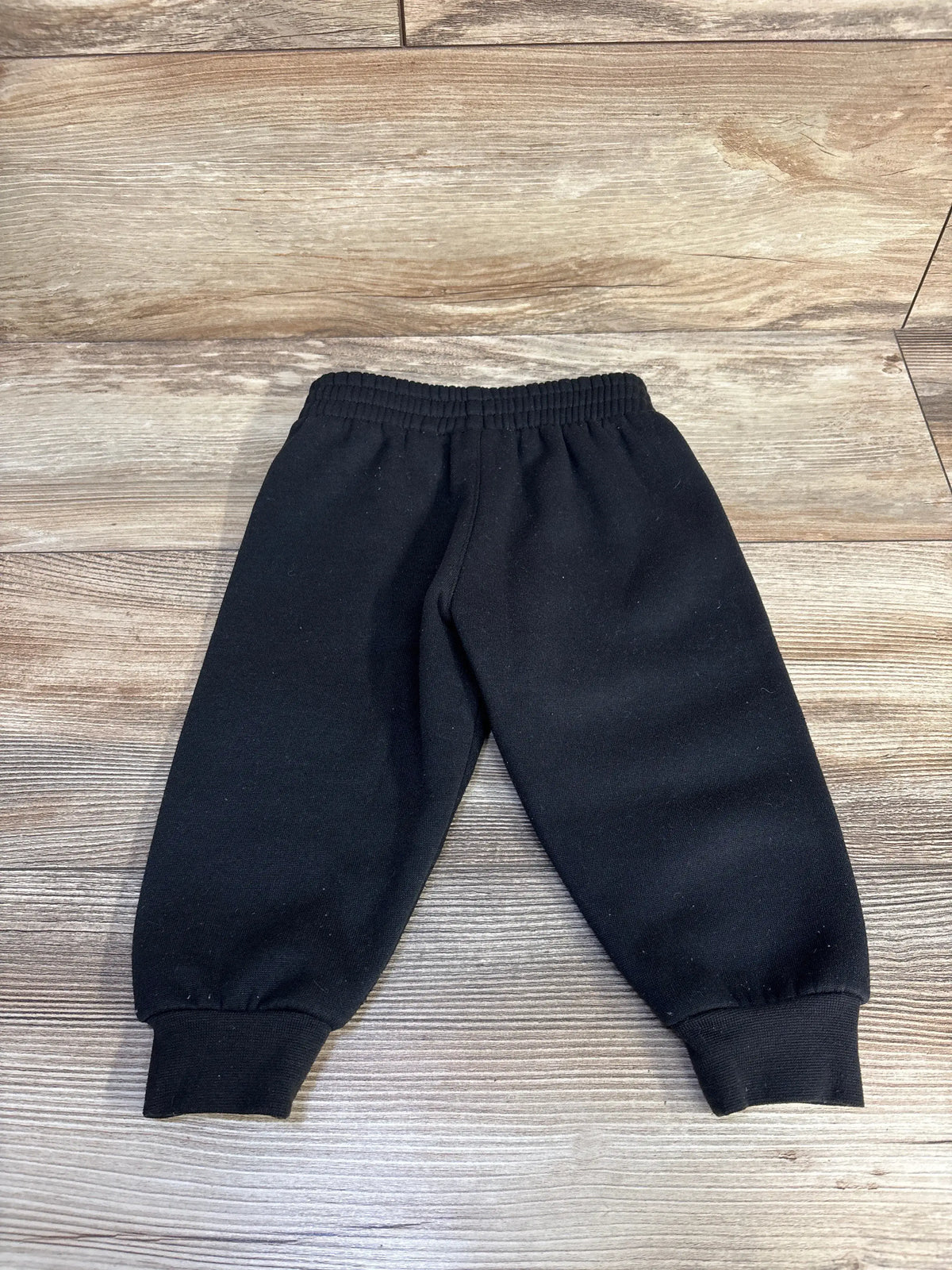 Front view of black Nike joggers, size 18 months, showcasing an elastic waist and cuffed ankles on a wood-look floor