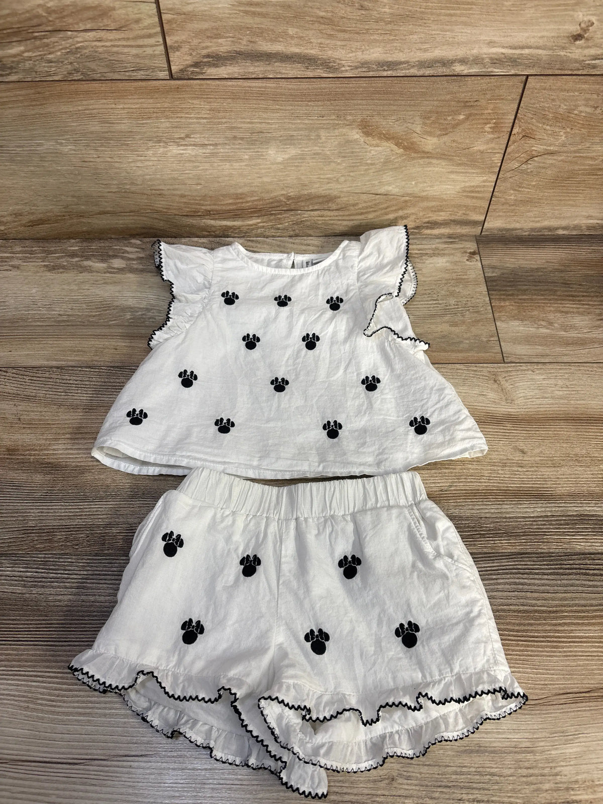 Flat lay of white Janie & Jack Disney Minnie two-piece set with black paw embroidery and ruffled black trim on wood floor