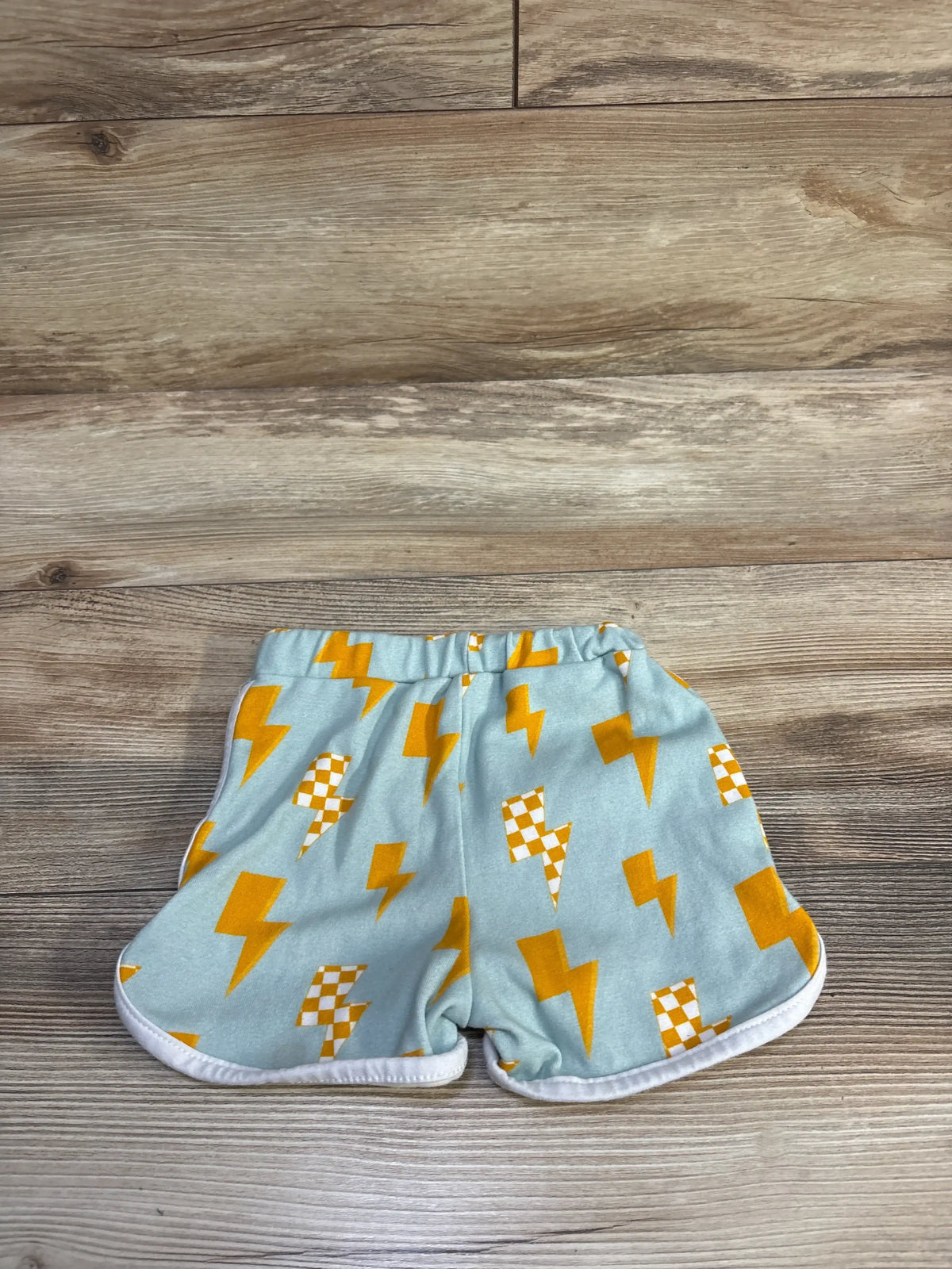 Flat-lay of Lucky Panda Kids blue lightning shorts with yellow and checkered lightning bolts, elastic waist and white trim on wood