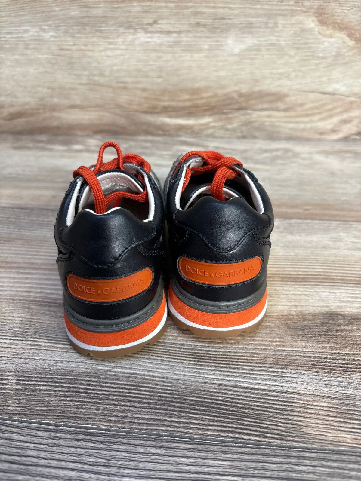 Rear view of Dolce and Gabbana Bambino black leather sneakers showcasing orange accents and red laces on a wood surface