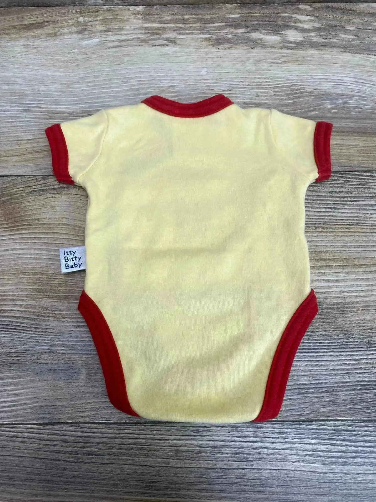 Flat lay of preemie kimono-style yellow baby bodysuit showcasing red trim and visible brand tag on wood background