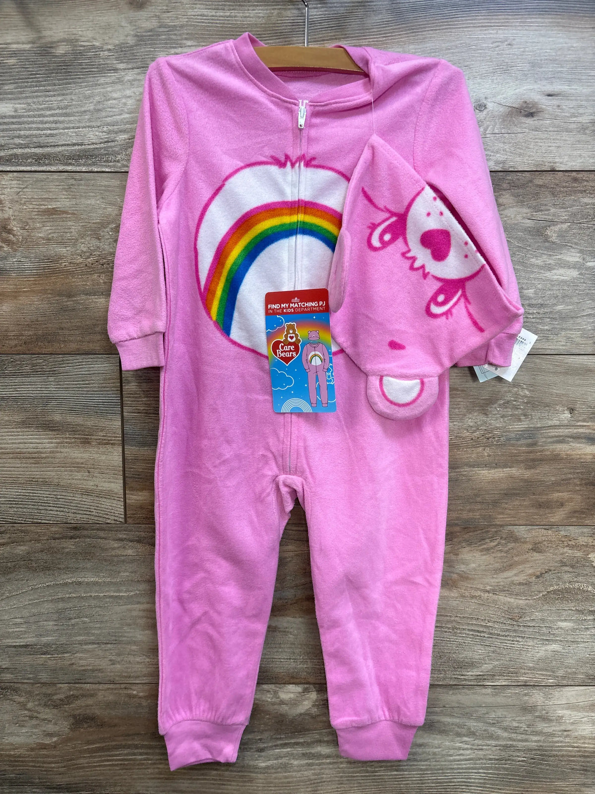 Front view of pink Care Bears zip sleeper with rainbow belly graphic and matching hat showing bear face on wood background