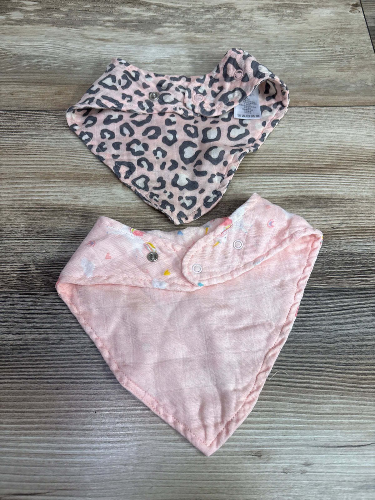 Front view of Angel Dear 2pk bandana bibs showcasing pink leopard and colorful patterns with snap closures on wood grain