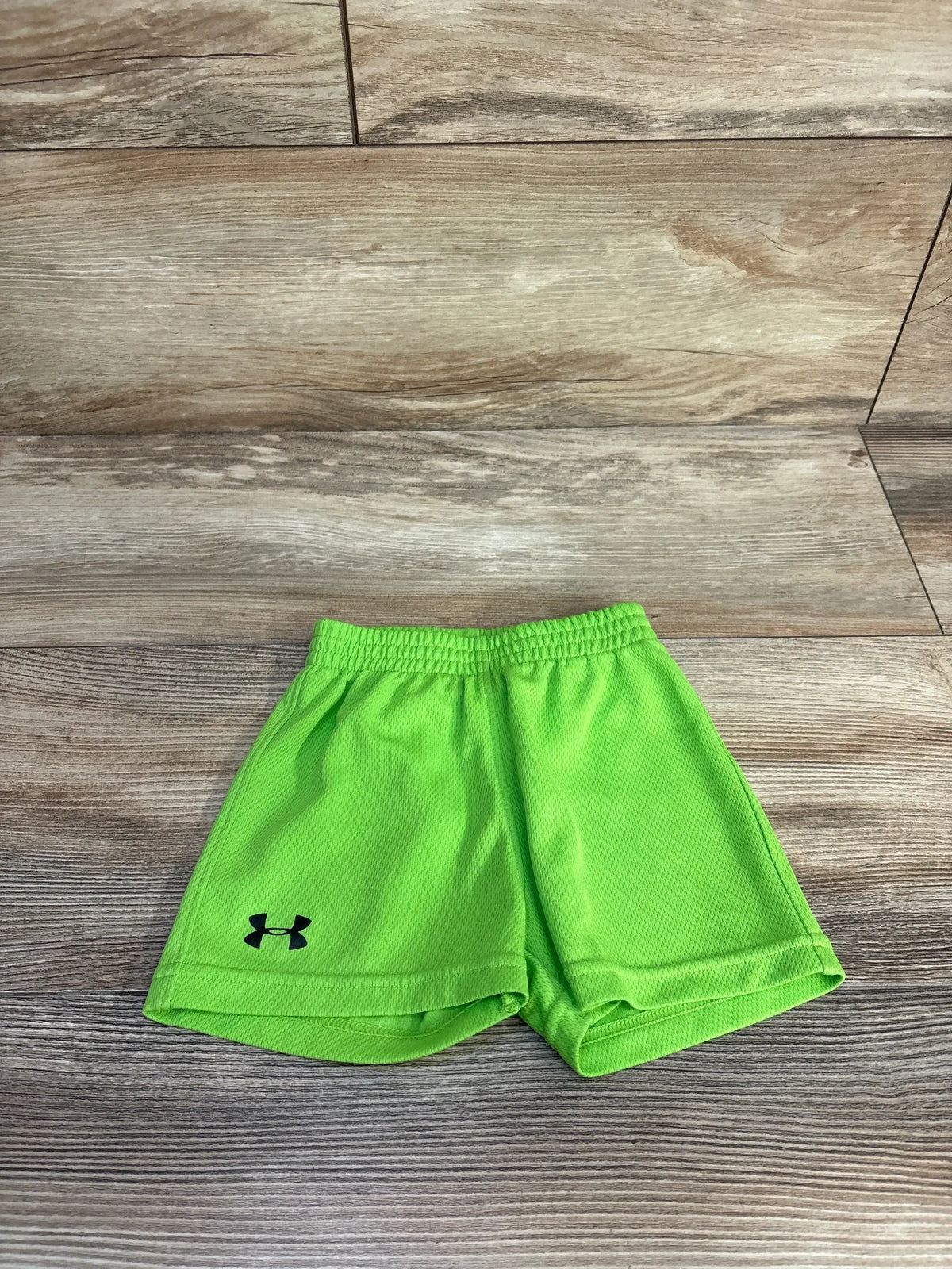 Front view of Under Armour mesh shorts in neon green, showcasing mesh fabric, elastic waistband, and black logo on a wood-look floor