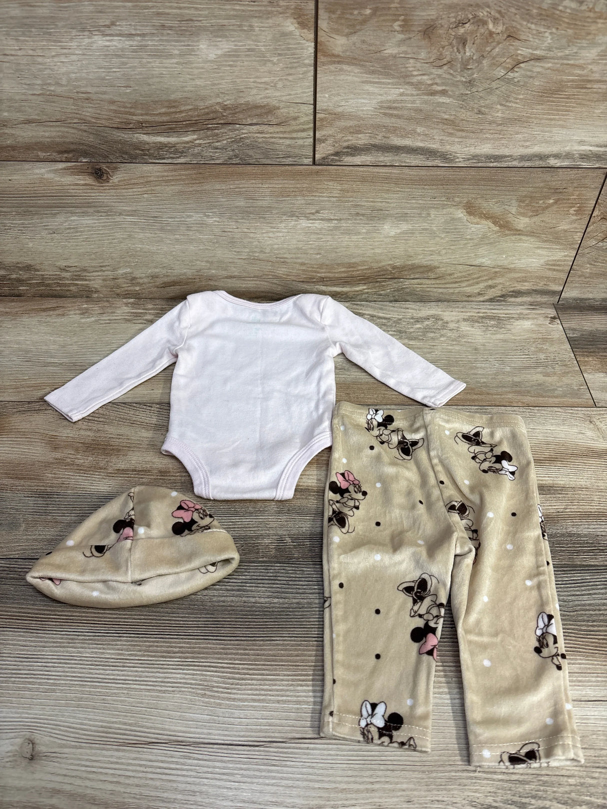 Flat lay of three piece Disney baby set with white long sleeve onesie and beige Minnie Mouse print pants and hat on wood plank floor