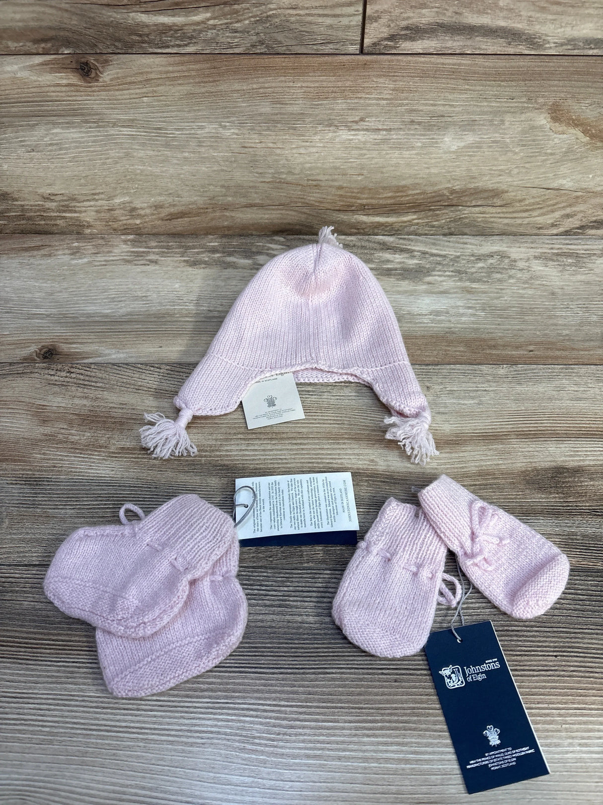 Pink knit baby hat, mittens, and booties set on wood background with Johnstons of Elgin tags.