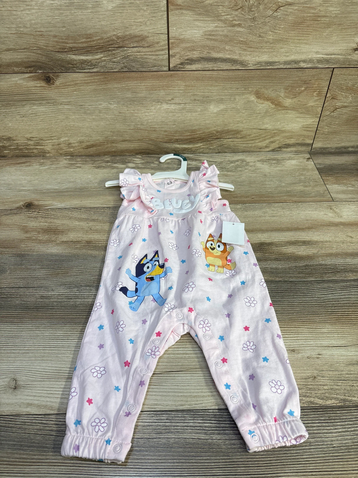 Front view of pink Bluey floral newborn jumpsuit with ruffle sleeves, character prints, and snap leg closures on wood floor