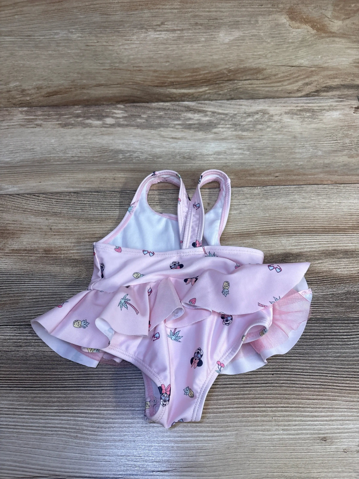Front view of pink Disney Baby Minnie Mouse one-piece swimsuit with ruffled tutu skirt on a wood-grain background