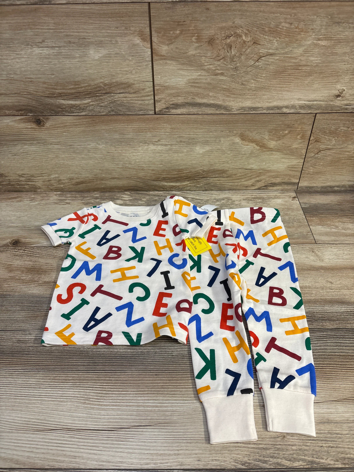 Flat-lay of Old Navy 2-piece ivory baby pajama set showing colorful scattered ABC letter print and cuffed ankle pants on wood plank floor