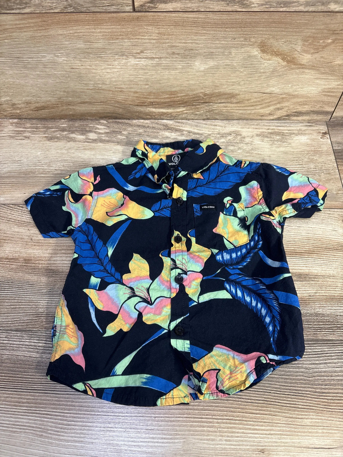 Front view of Volcom navy button-up shirt in size 2T showcasing colorful floral and leaf print with short sleeves and chest pocket on a wood floor