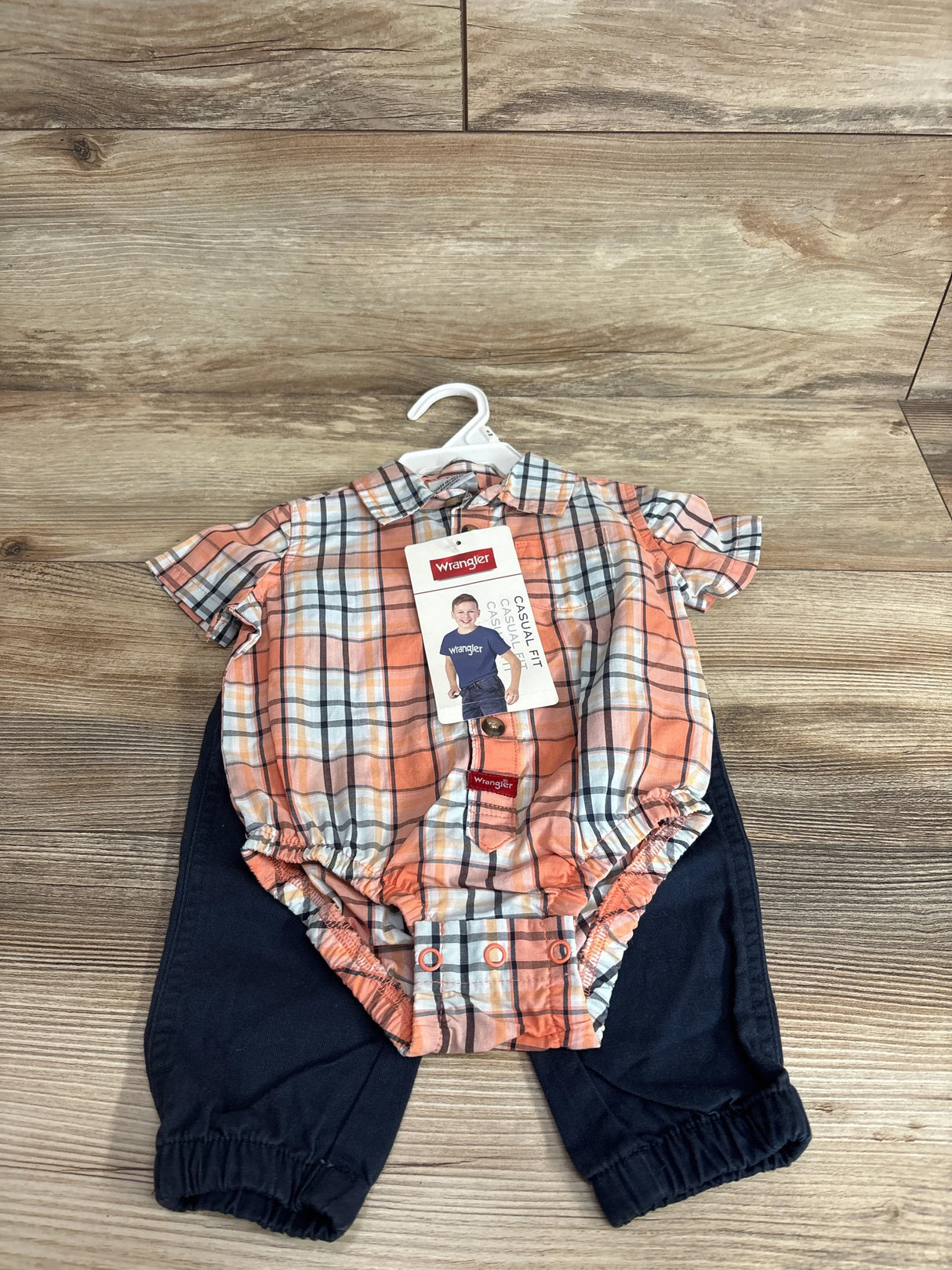 Top-down view of Wrangler orange plaid short-sleeve bubble romper with snap crotch and collar, paired with navy pants on wood floor