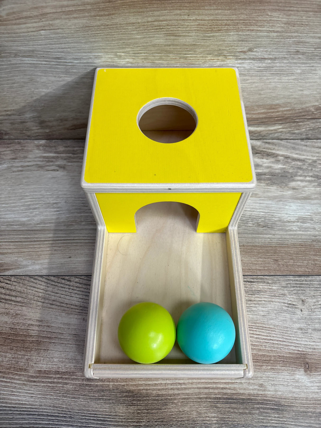 Wooden Montessori toy with yellow box and two balls, green and blue, on wood floor