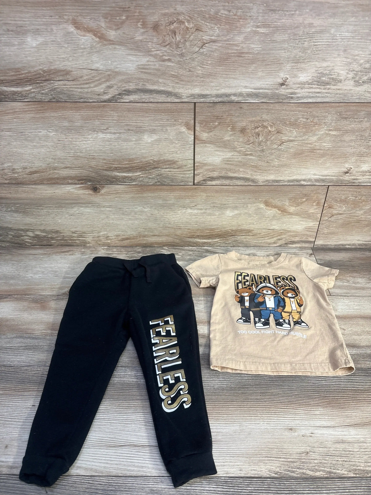 Top-down view of Fashion Nova Contact Mini Fearless Squad jogger set with beige graphic tee and black pants on wood floor