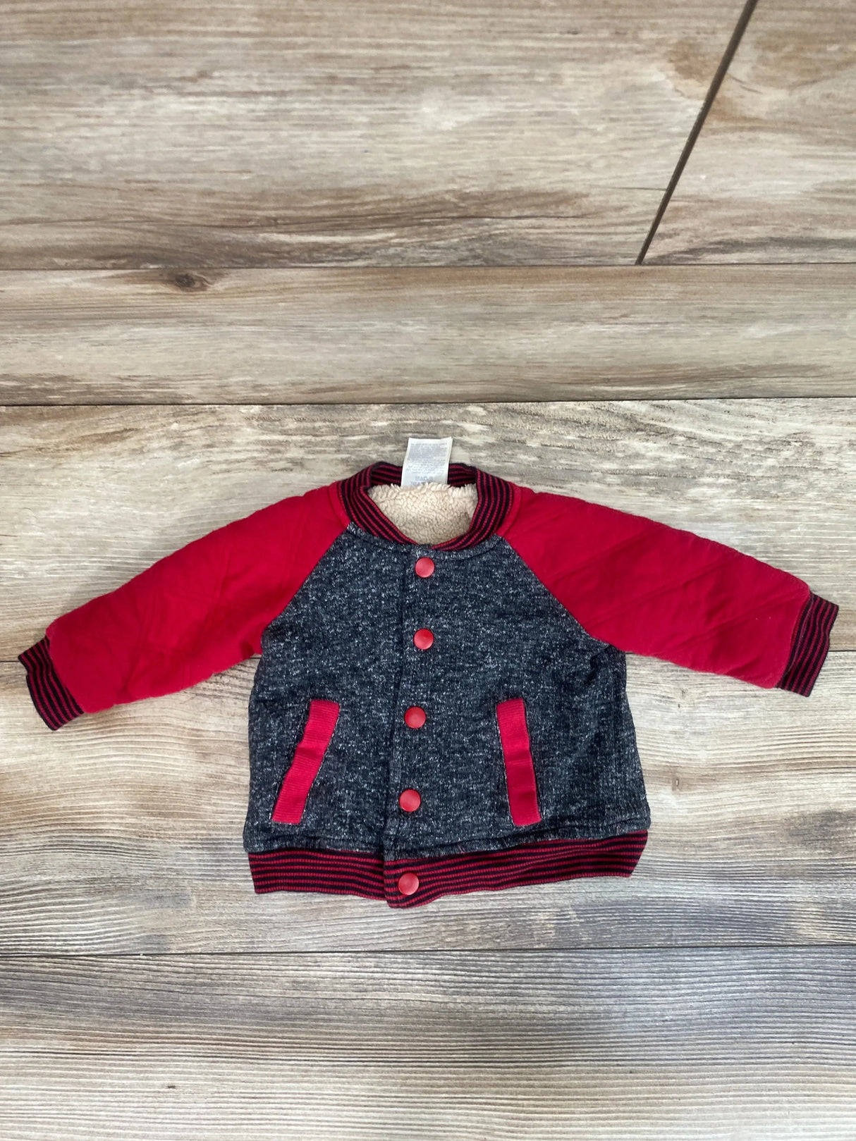 Flat-lay of 3-6 month baby jacket with red sleeves, marled gray body, red snap buttons and striped ribbed cuffs on wood floor