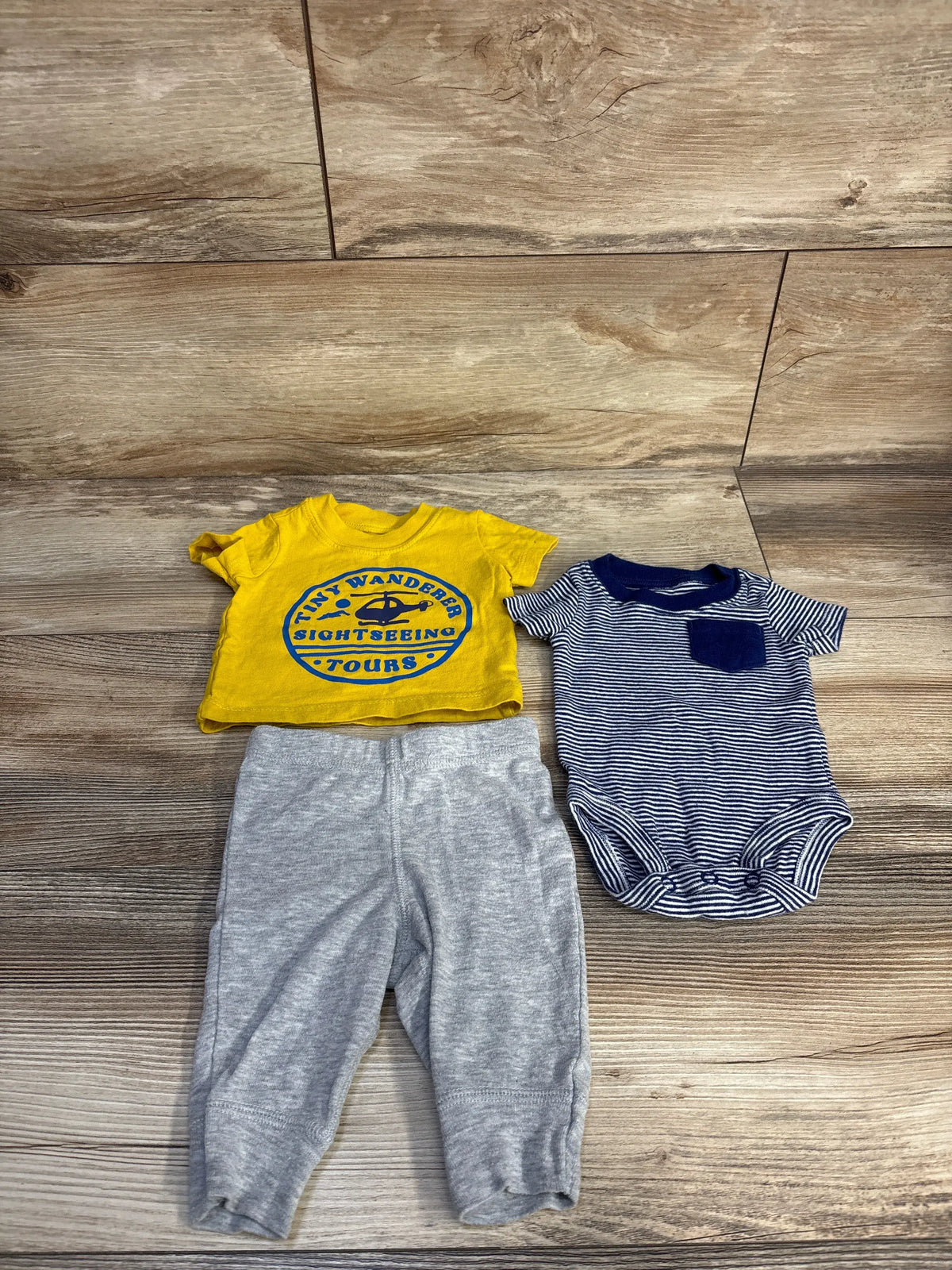 Top-down view of Carter’s newborn three-piece set showcasing yellow shirt, striped bodysuit, and gray pants on wood flooring