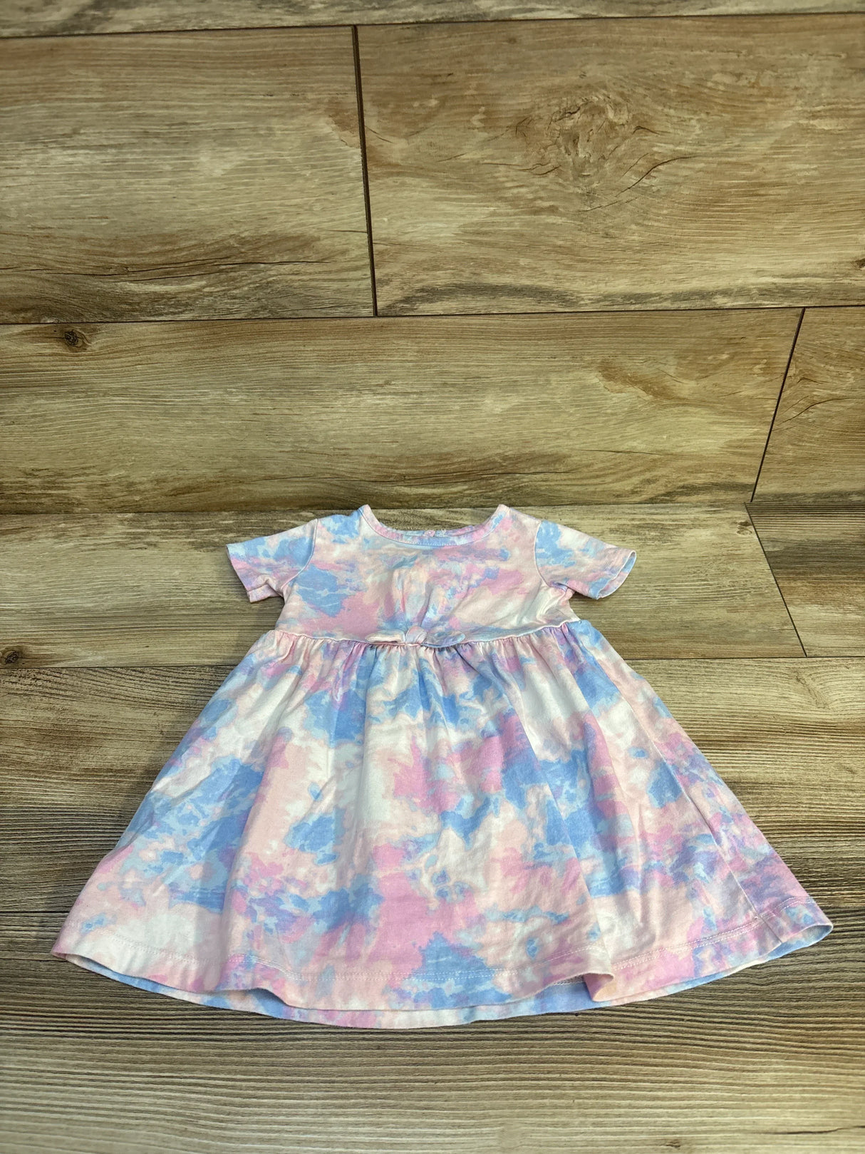 Flat lay of Carter's short-sleeve baby dress showing pink and blue tie-dye pattern and gathered waist skirt on wood plank background