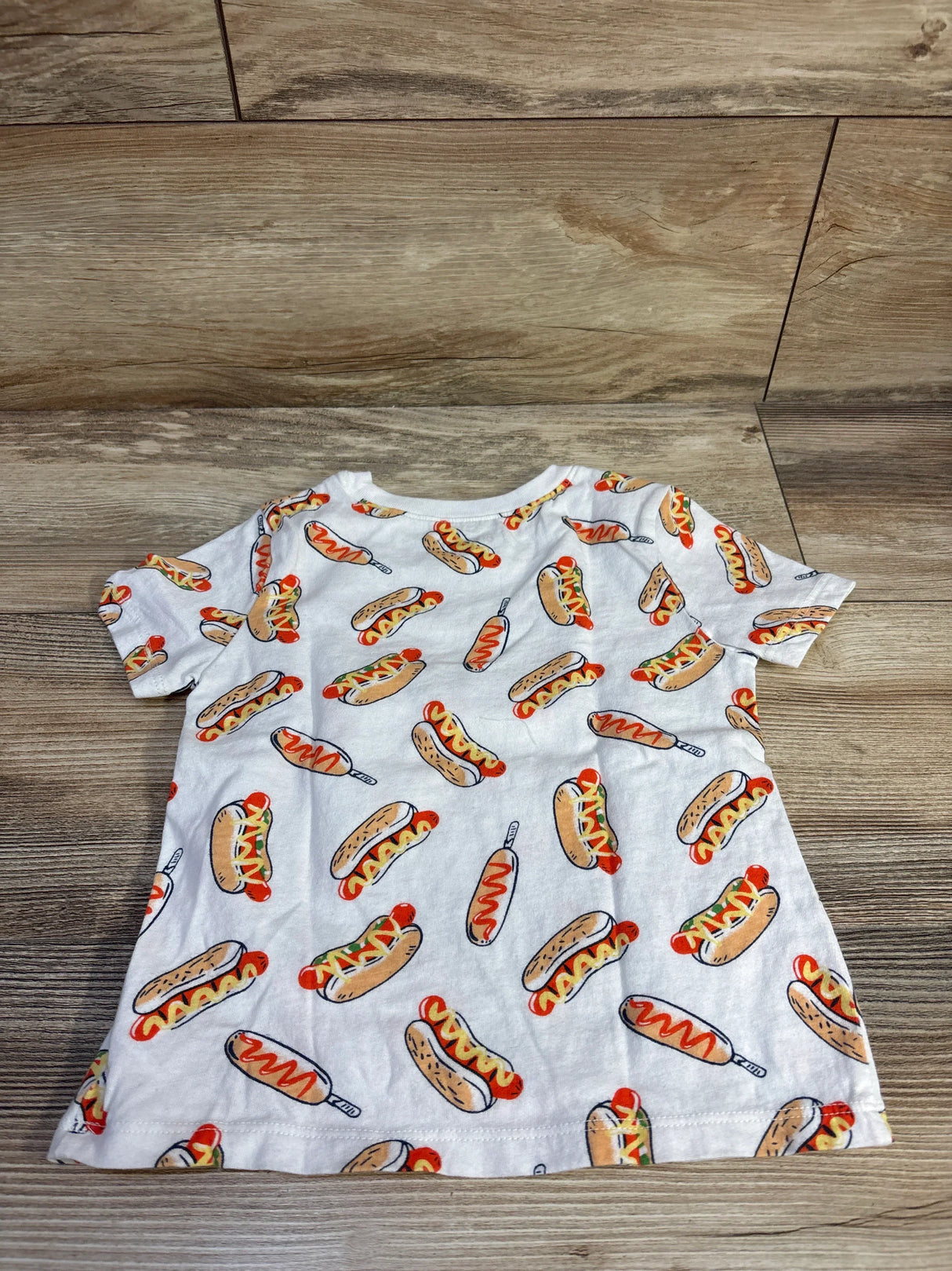 Flat-lay of white Old Navy 4T short-sleeve toddler shirt showcasing all-over hot dog and corn dog print on wood floor background