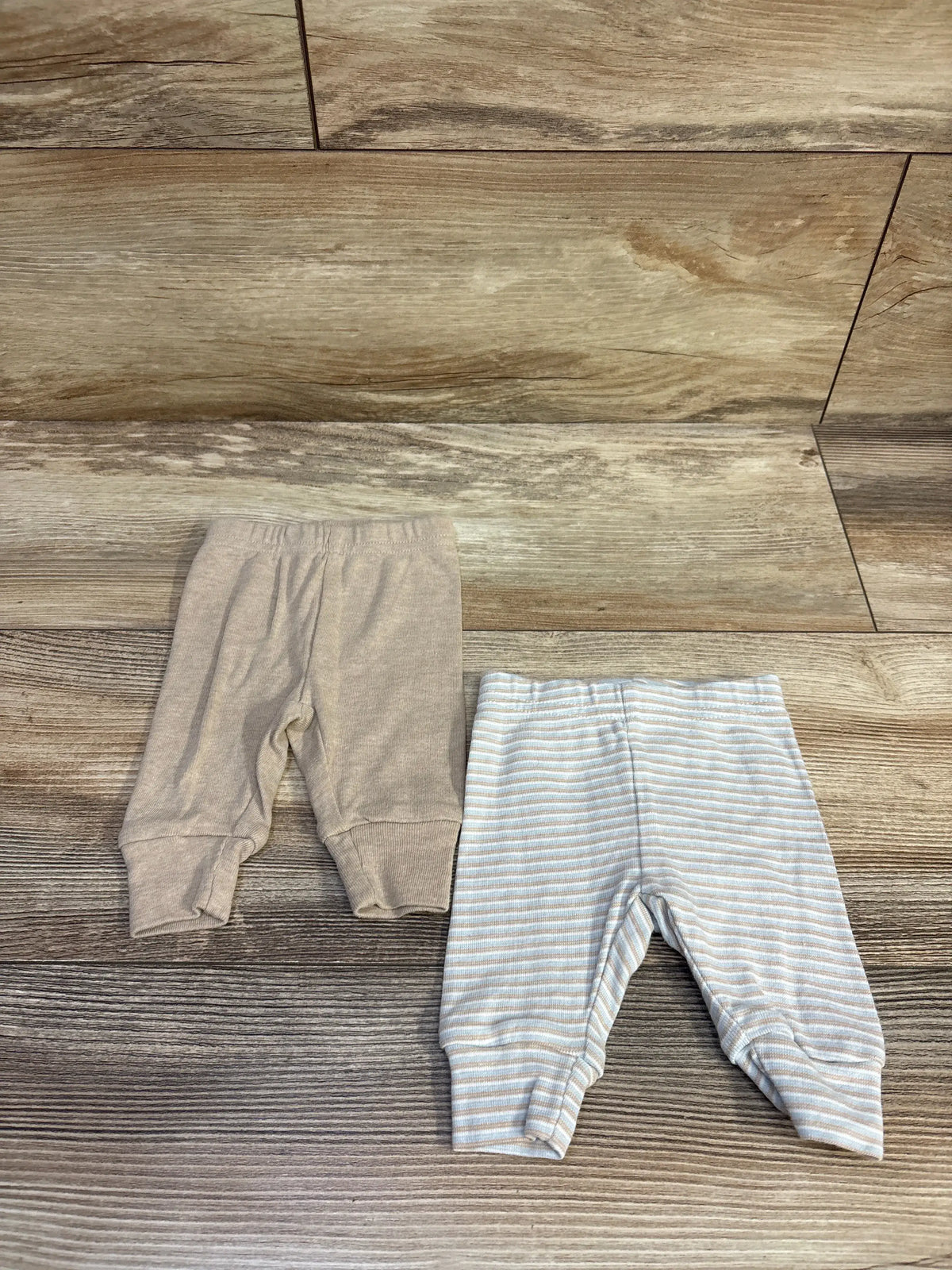 Top view of Just One You two-pack newborn pants showcasing beige and blue-and-beige striped pairs on a wood-look floor
