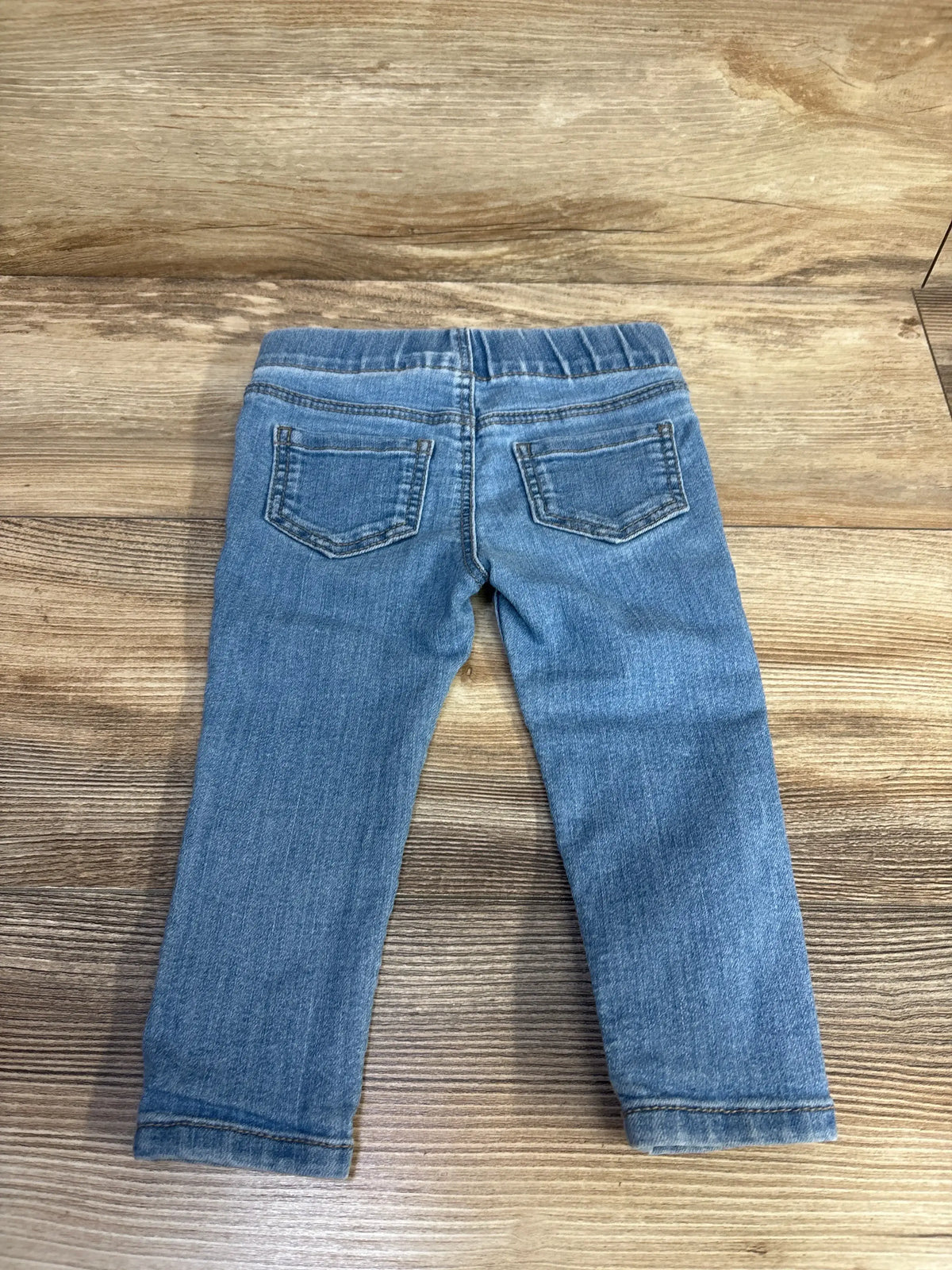 Rear view of blue Oshkosh jeggings pants in size 18 months showcasing two back pockets on a wood background