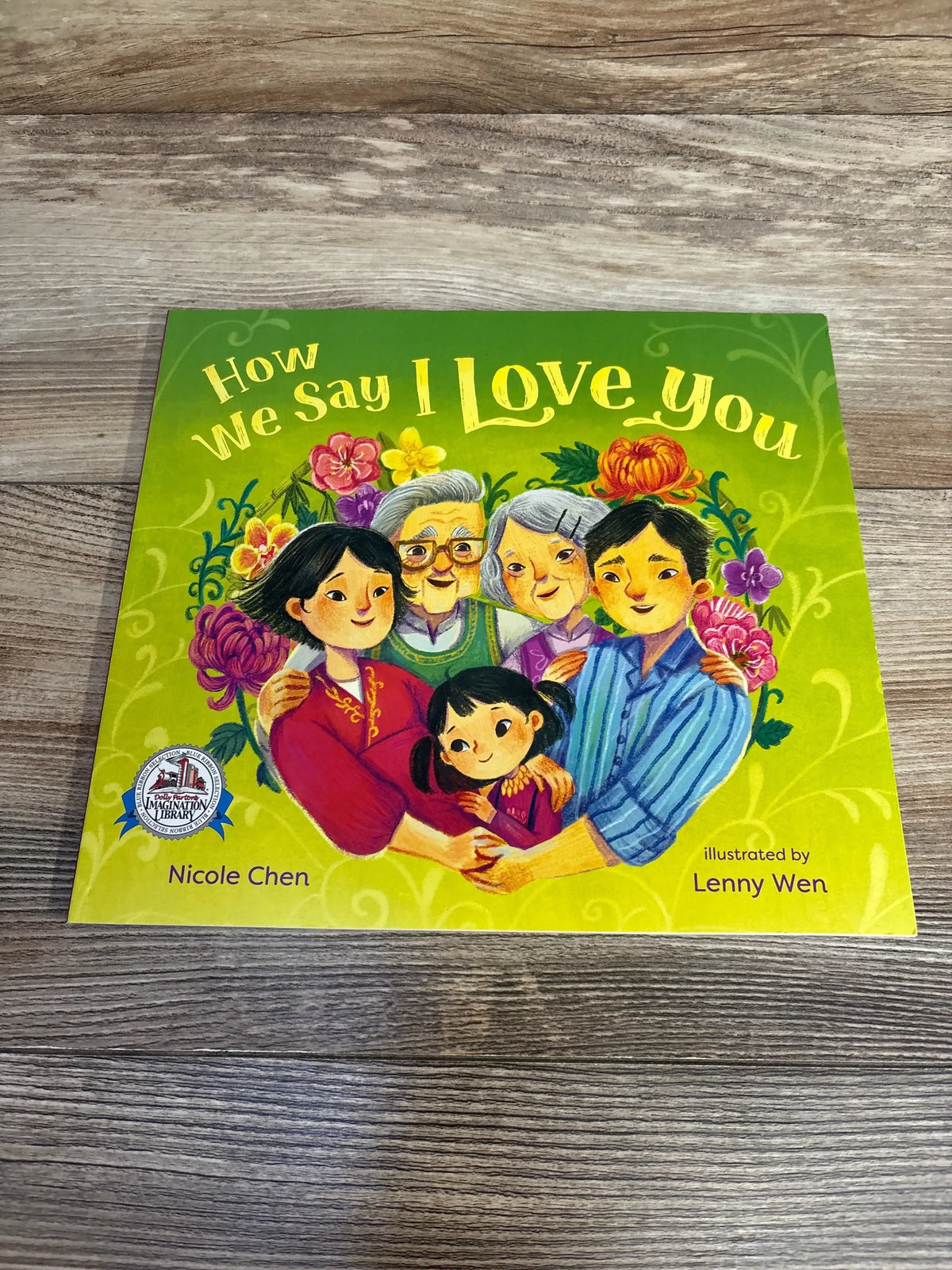 Flat-lay front cover of How We Say I Love You paperback showcasing colorful family illustration and title with wood tabletop background