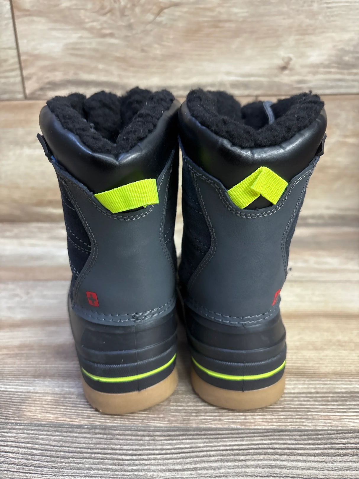 Rear view of Swiss Tech kids’ black winter boots showing fleece lining, neon yellow pull tabs, and rubber soles on wood floor background