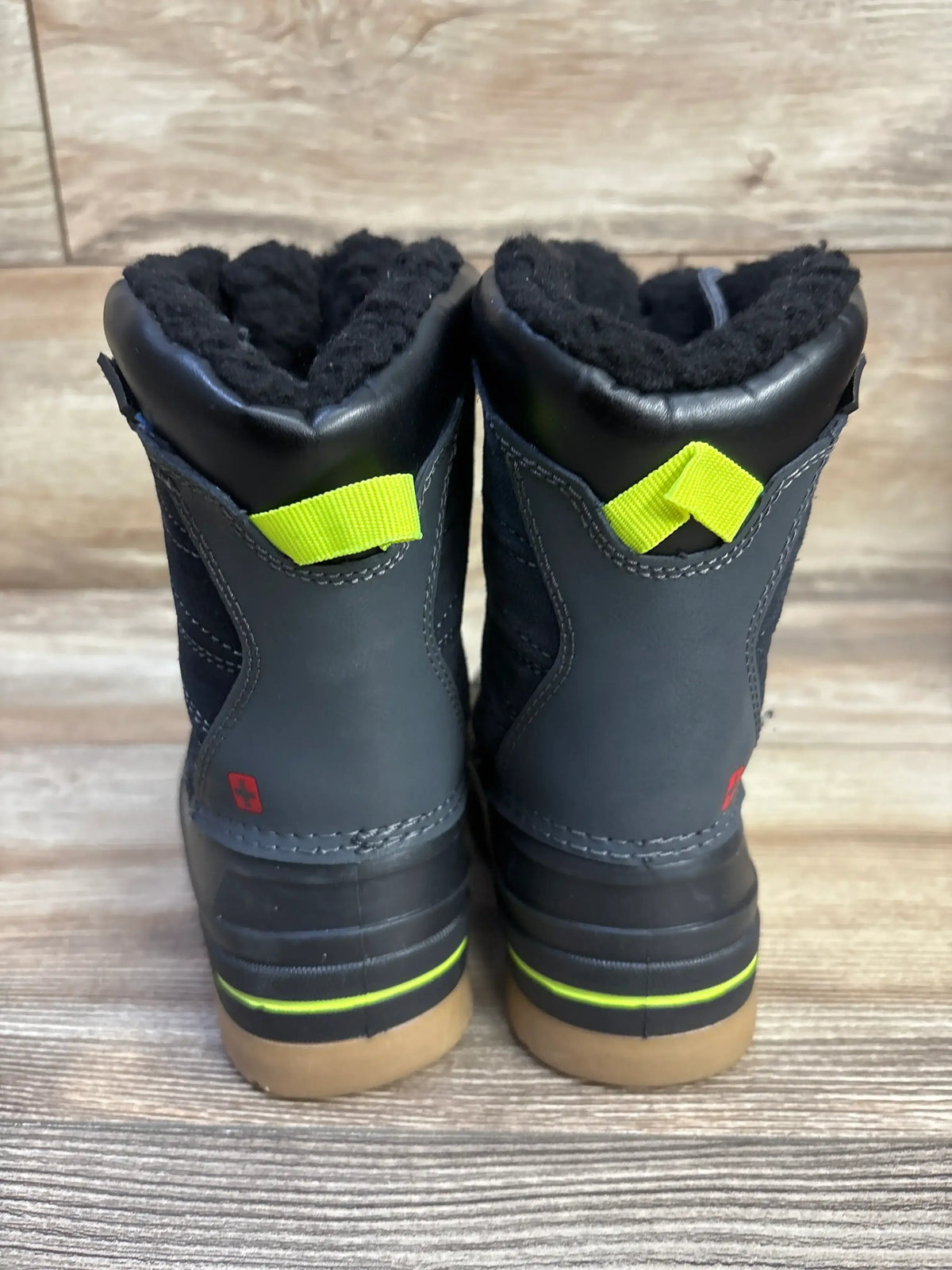 Rear view of Swiss Tech kids’ black winter boots showing fleece lining, neon yellow pull tabs, and rubber soles on wood floor background