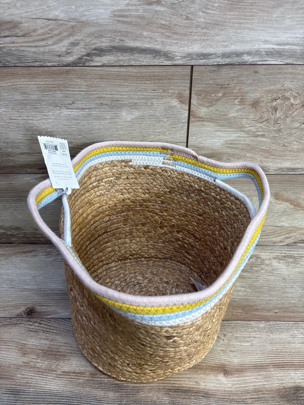 Top-down view of Cloud Island natural woven round storage bin showcasing braided seagrass texture and pastel pink, yellow, and blue rim with handles on wood floor