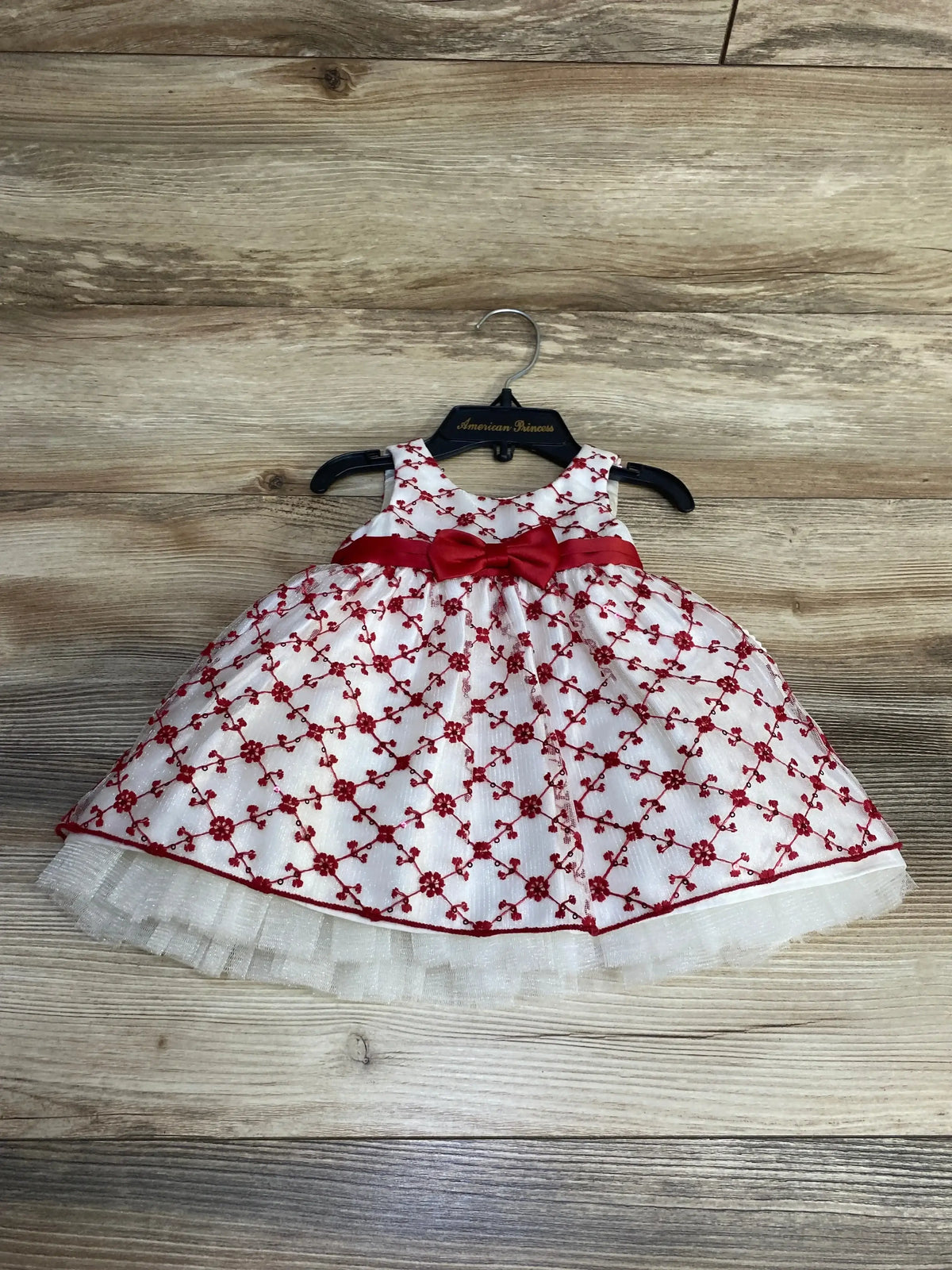 Front view of white sleeveless baby dress with red floral embroidery and red waist bow, tulle skirt on hanger on wood