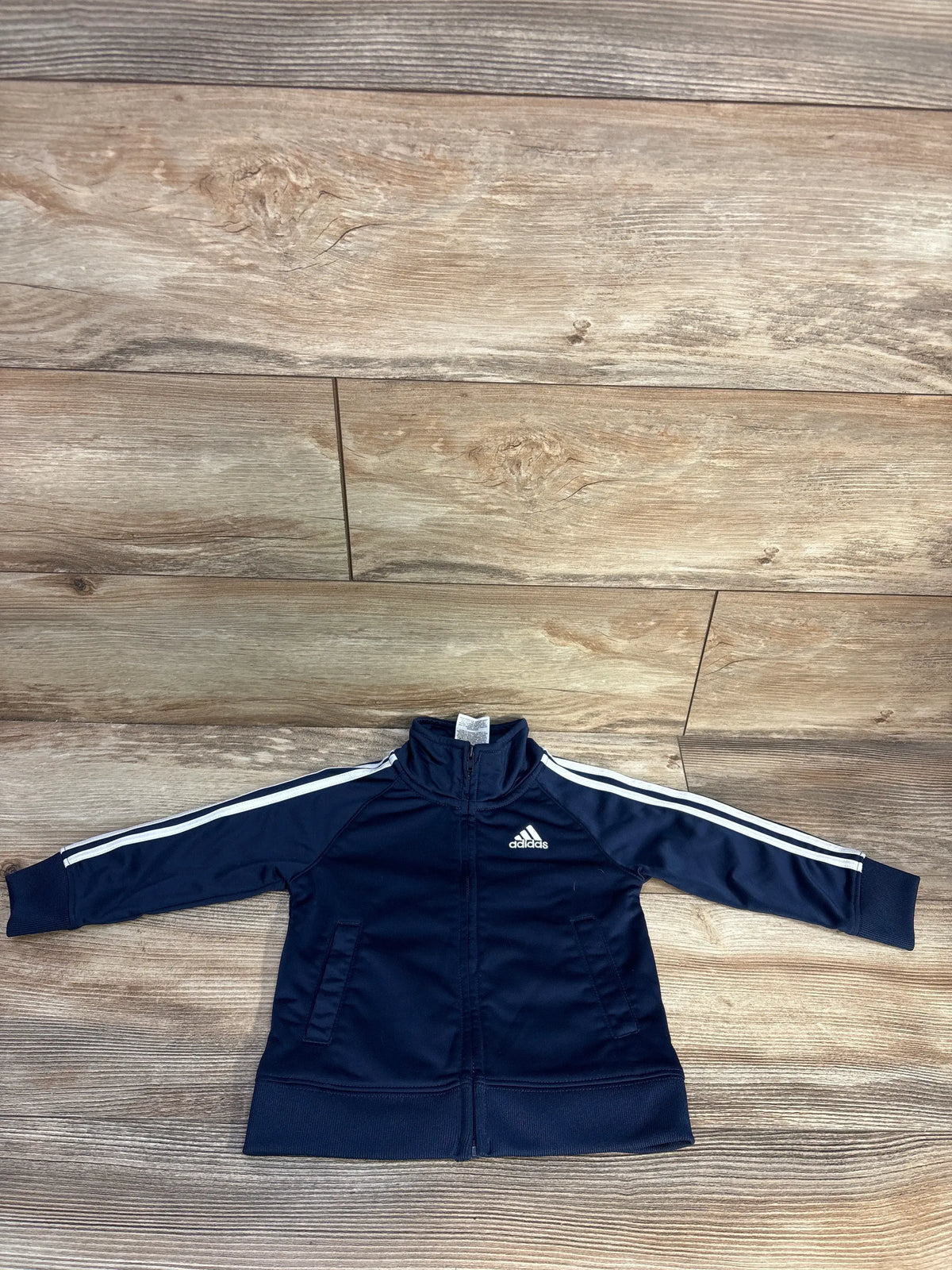 Flat lay front view of navy Adidas track jacket with full zip and white sleeve stripes on wood background, size 18 months