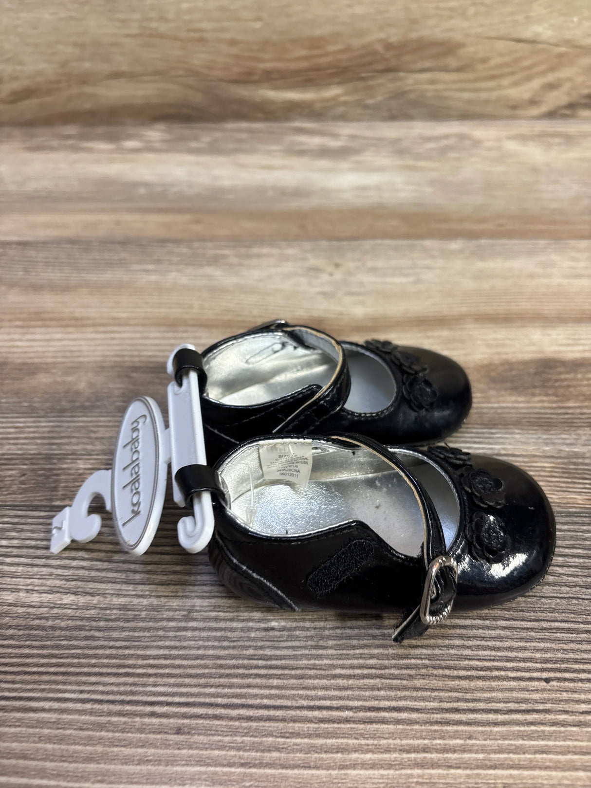 Top view of Koala Baby black Mary Jane flats showing floral toe detail, ankle strap with buckle and Velcro on wood background