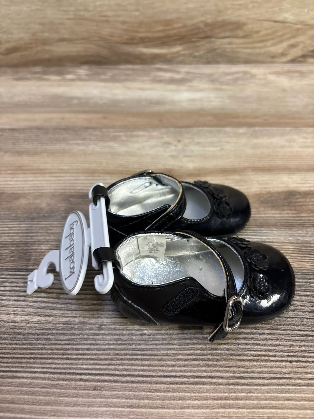 Top view of Koala Baby black Mary Jane flats showing floral toe detail, ankle strap with buckle and Velcro on wood background