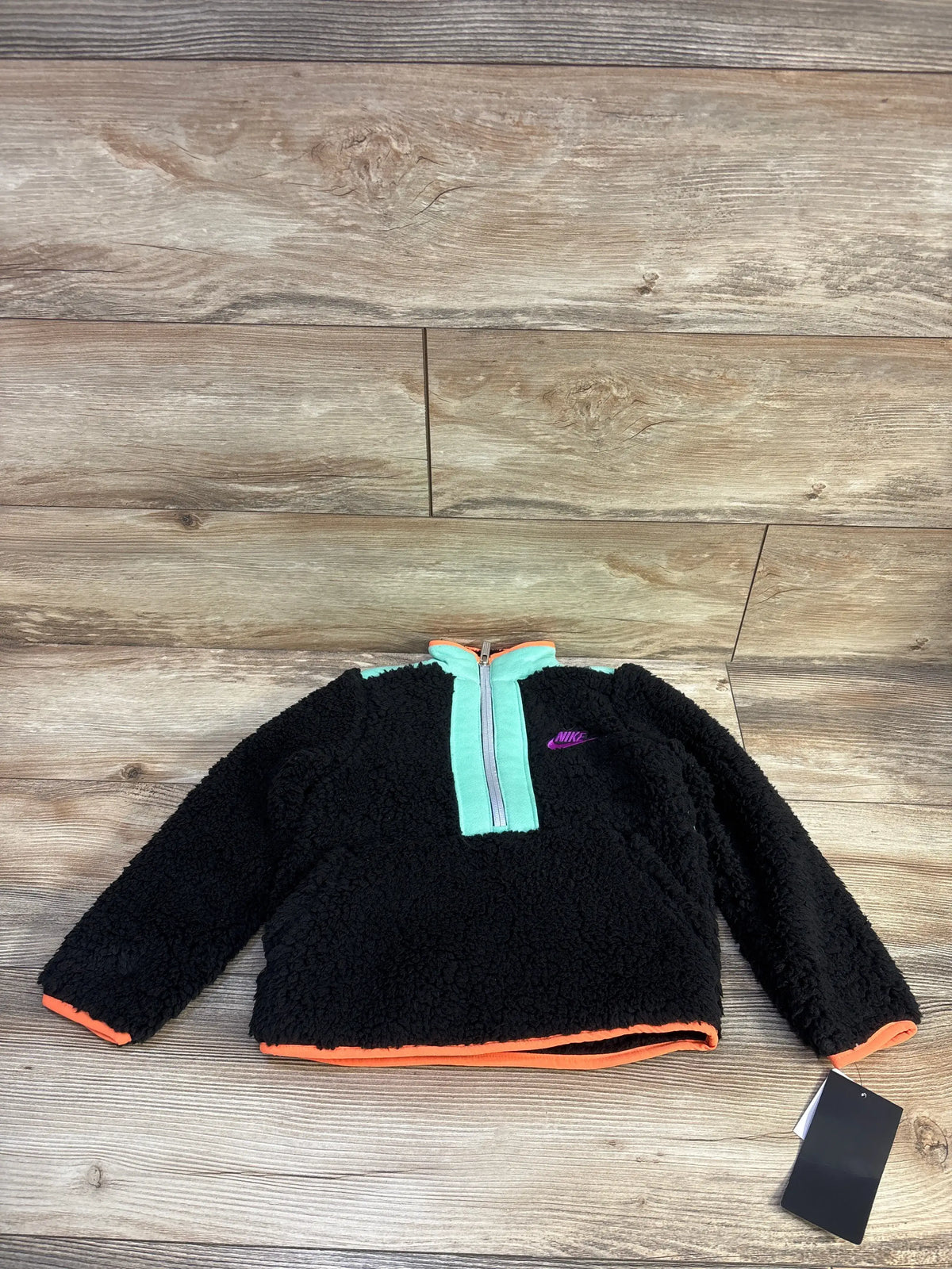 Front view of Nike Illuminate Sherpa jacket, toddler 4T, showing black sherpa fleece, mint half-zip placket, purple Nike logo and orange trim on wooden floor