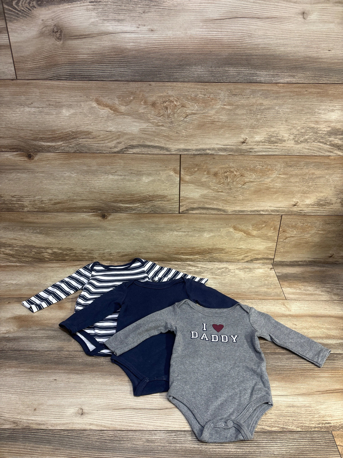 Front view of Hudson Baby 3-pack long-sleeve bodysuits in navy, striped, and gray, with I love Daddy text on wood background