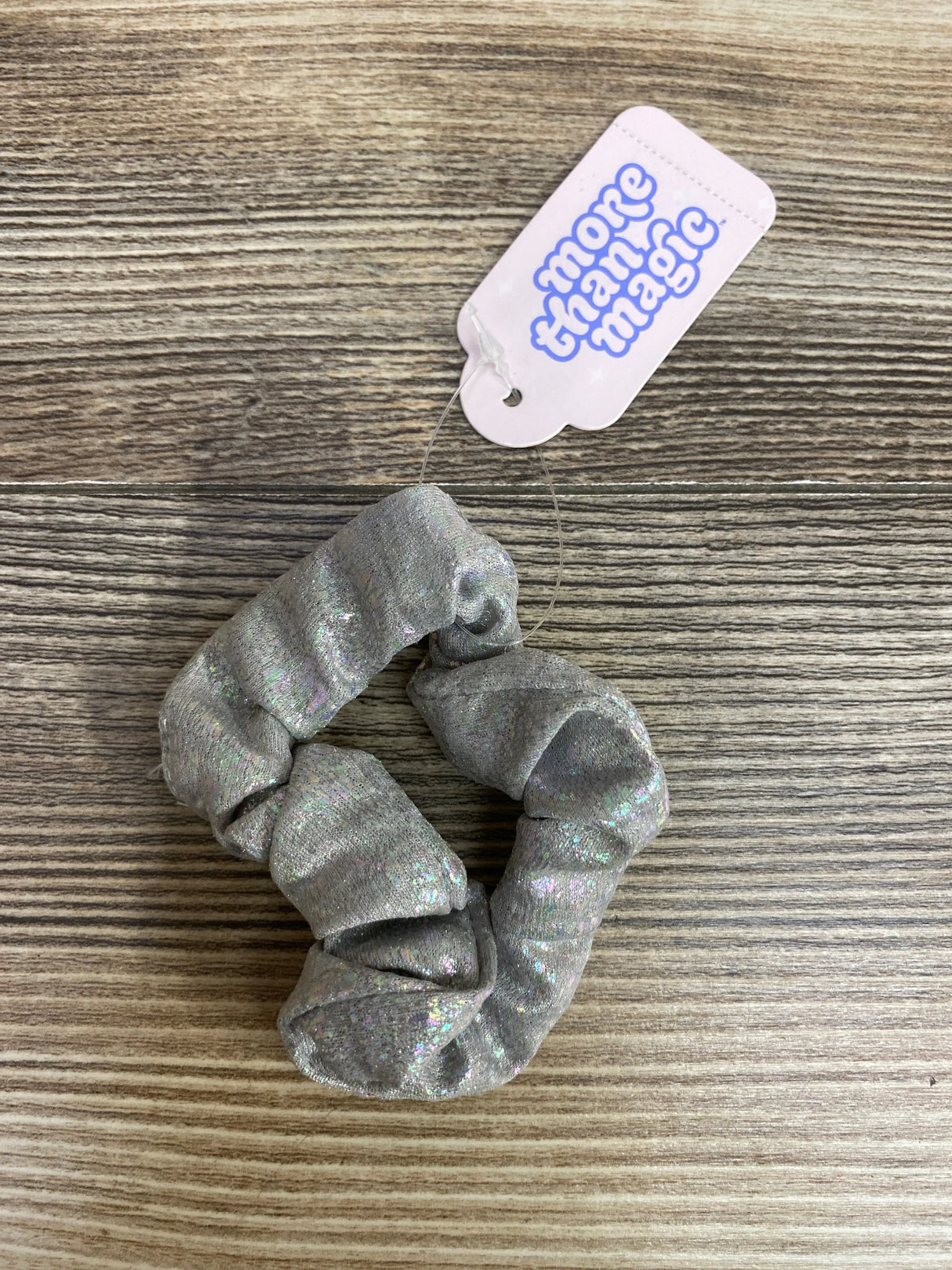 Silver glittery fabric scrunchie with 'more than magic' tag on wooden surface