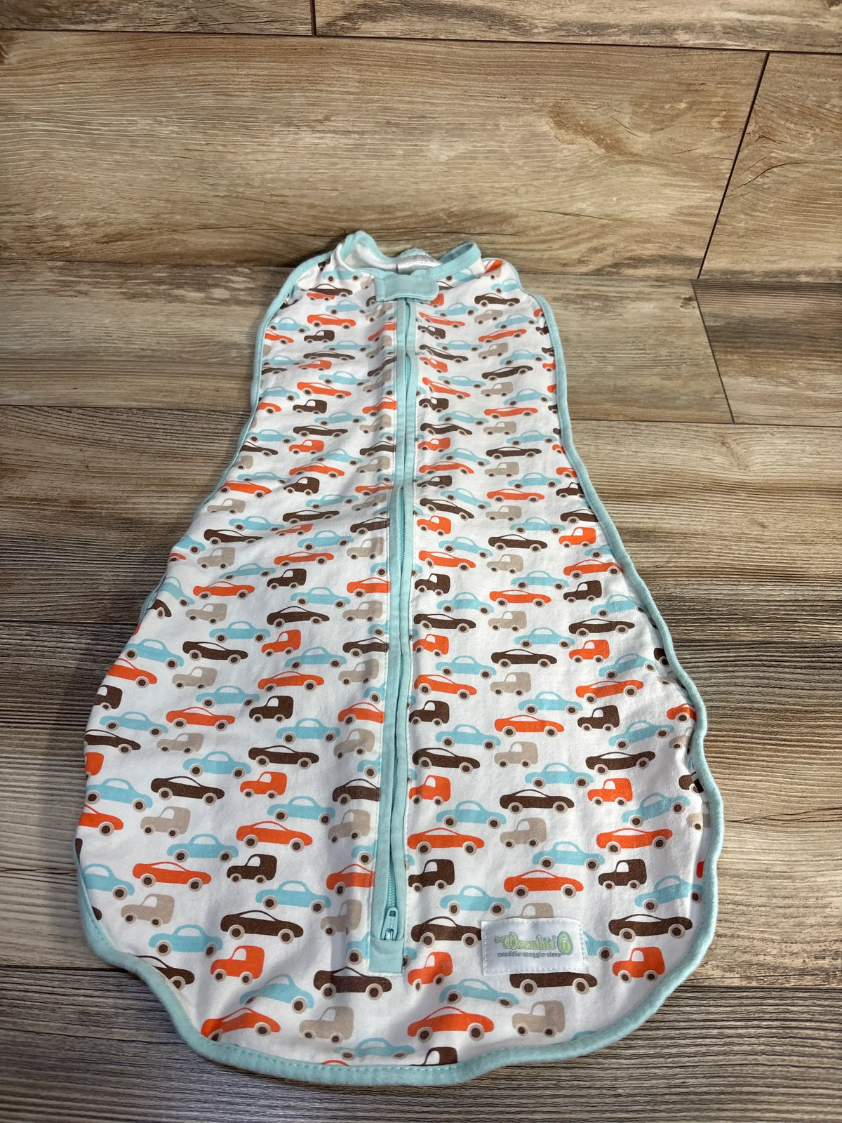 Front view of the Woombie Original Swaddle in white car print, showcasing a two-way zipper and turquoise trim on a wood floor