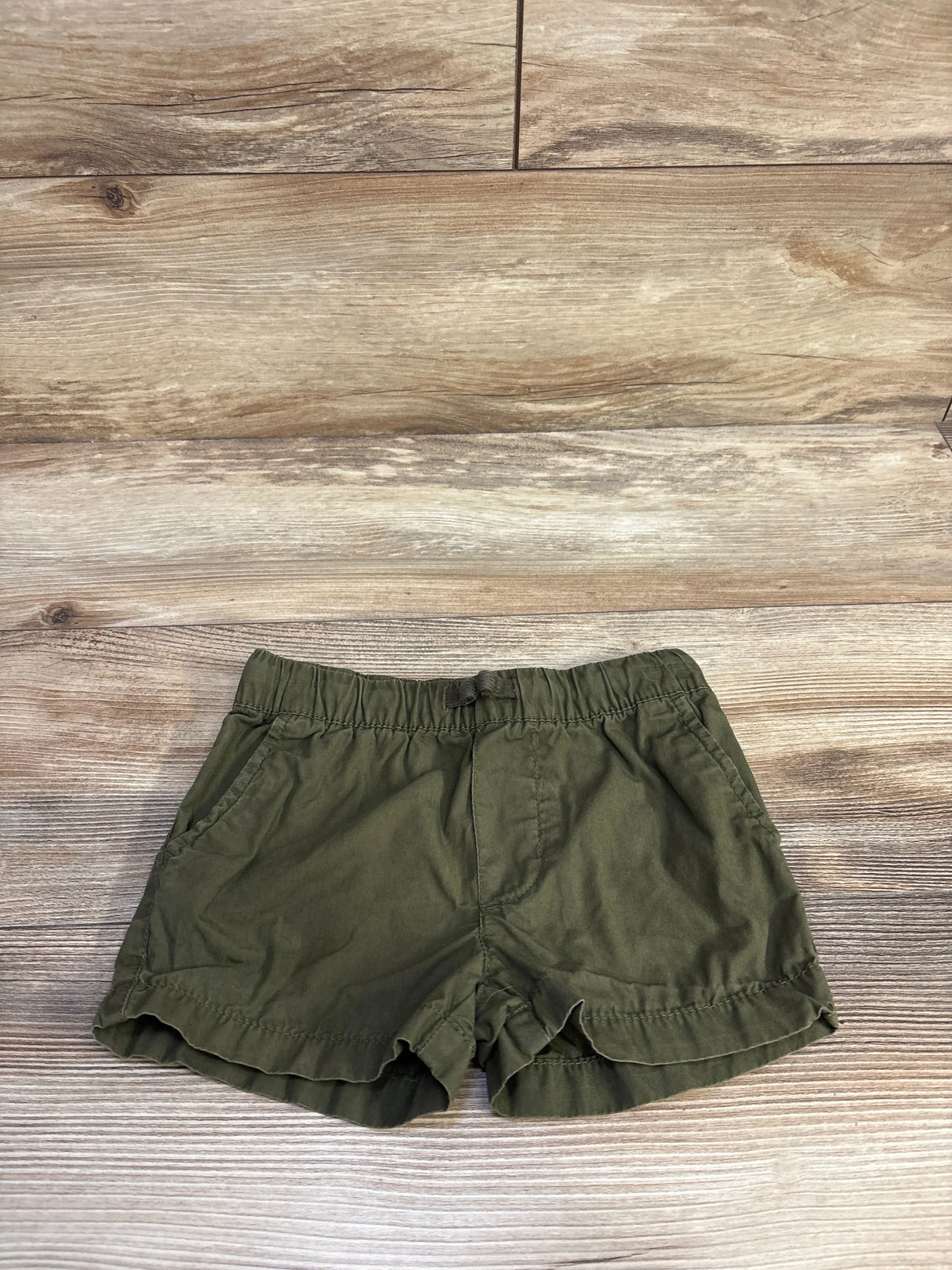 Top-down view of Carter’s green toddler drawstring shorts with elastic waistband and scalloped hem on wood background