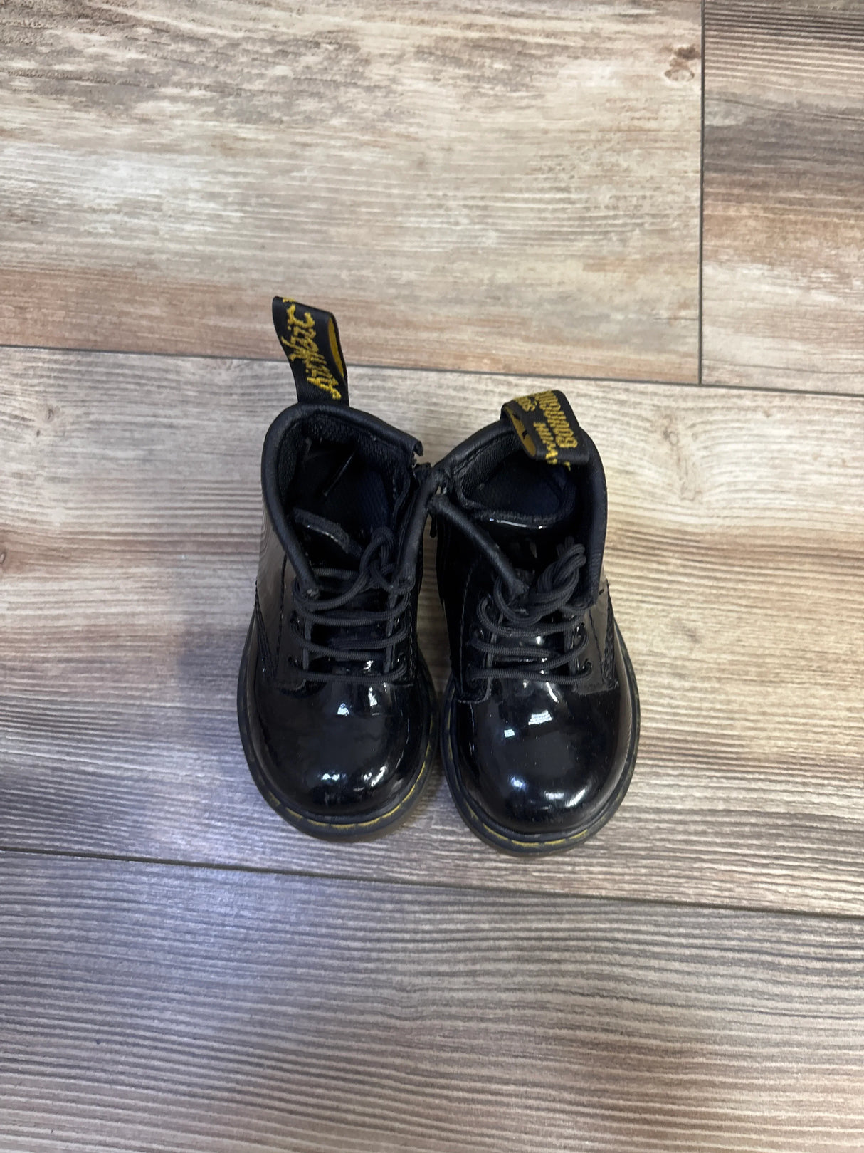 Overhead view of toddler black patent leather Dr Martens boots with laces, yellow welt stitching and heel loops on wood floor