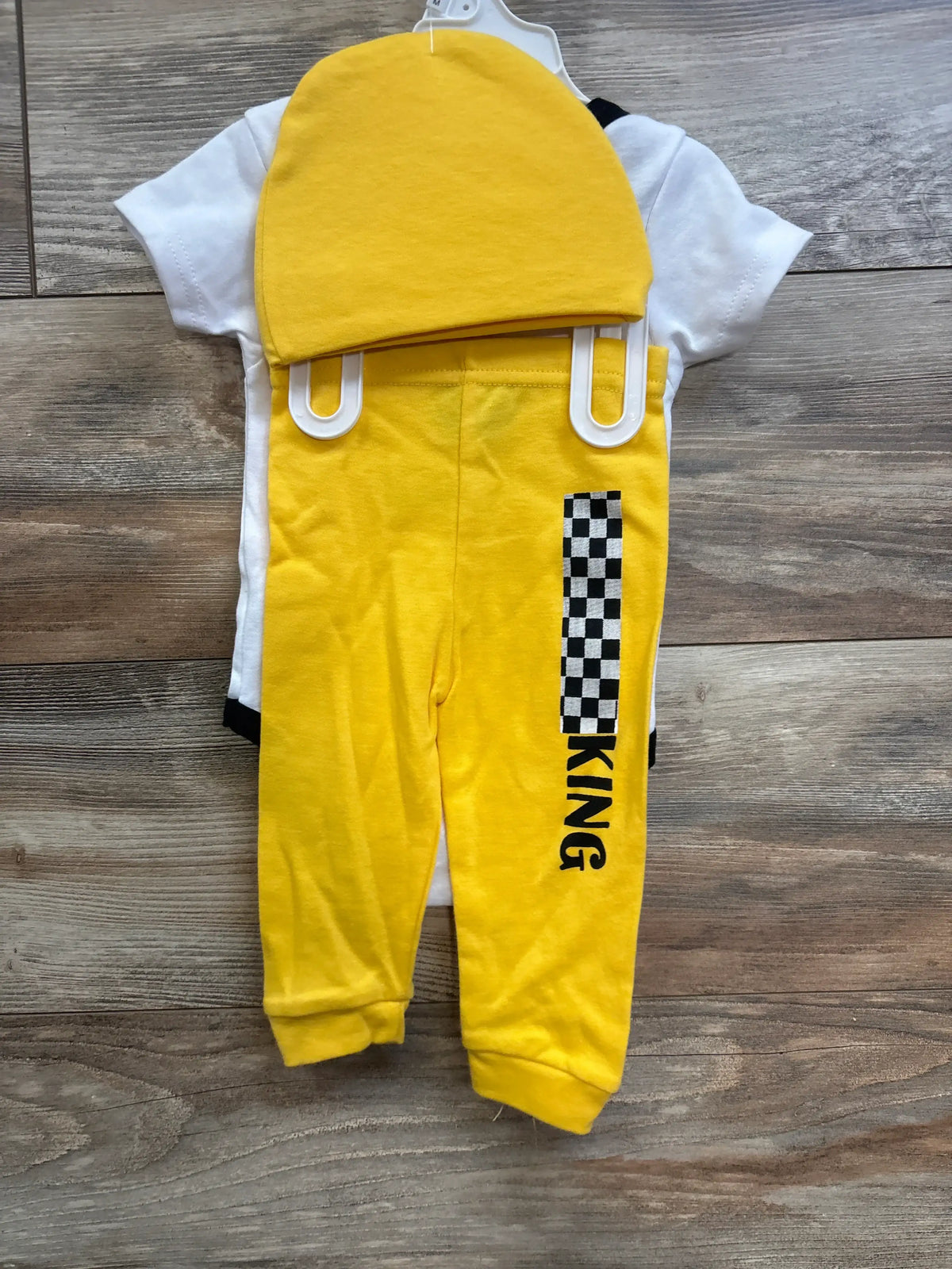 Flat lay of white bodysuit, yellow pants, and matching yellow hat; pants show checker stripe and KING text on wood floor